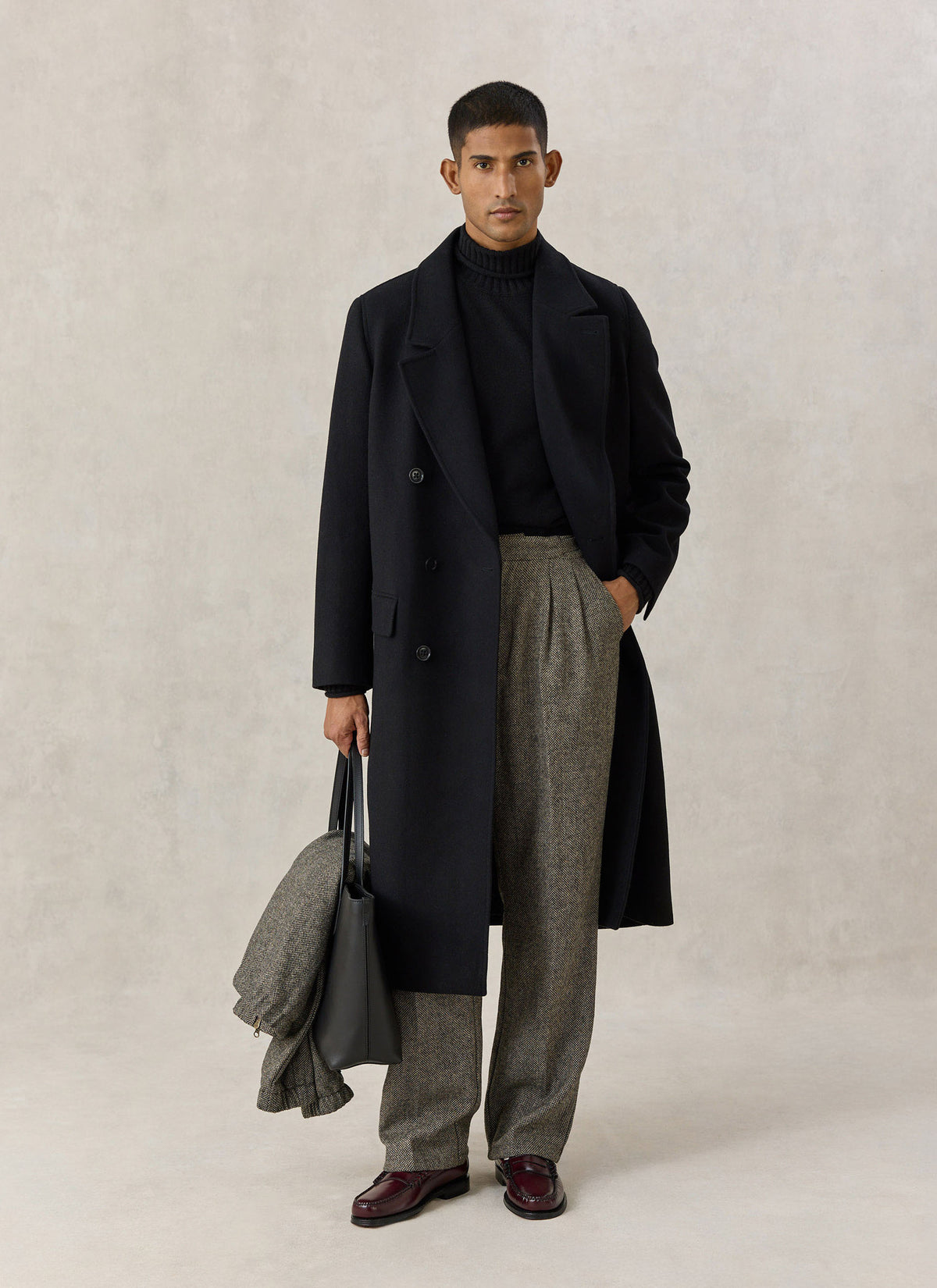 A model wearing the Percival black wool blend double-breasted overcoat with trousers, a dark knit, and holding a bag.