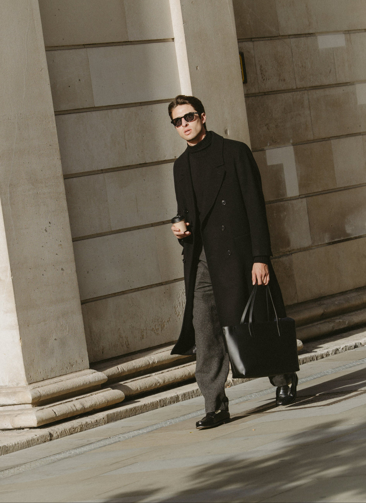 A model wearing a black wool blend double-breasted overcoat, dark turtleneck, and charcoal trousers while walking outdoors.