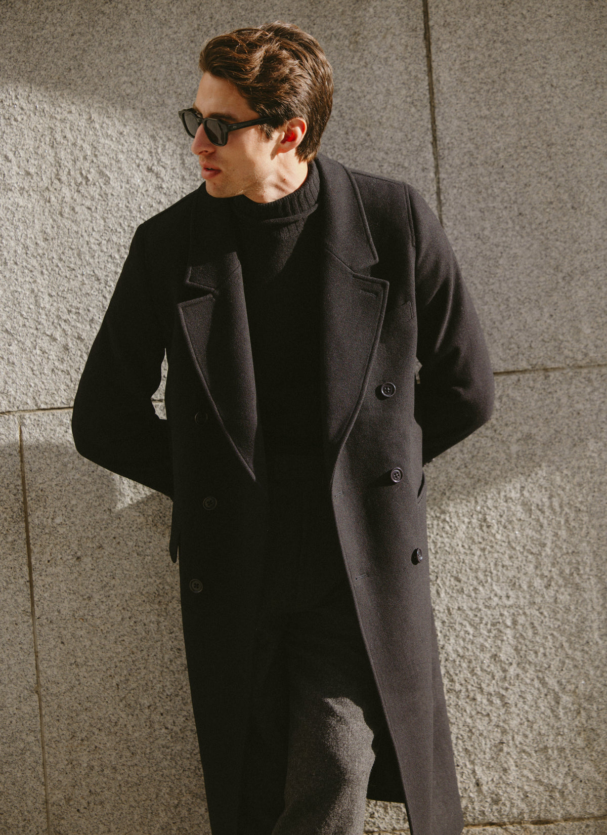 A model wearing a black wool blend double-breasted overcoat and dark turtleneck against a stone wall.