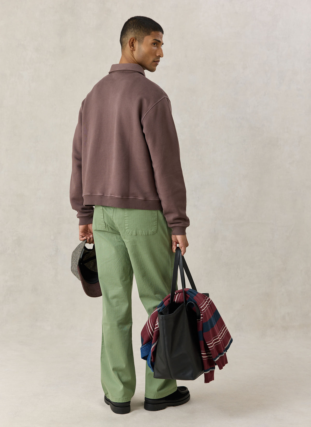A model from the back in Percival brown Dudley sweatshirt, green trousers, holding a tote bag.