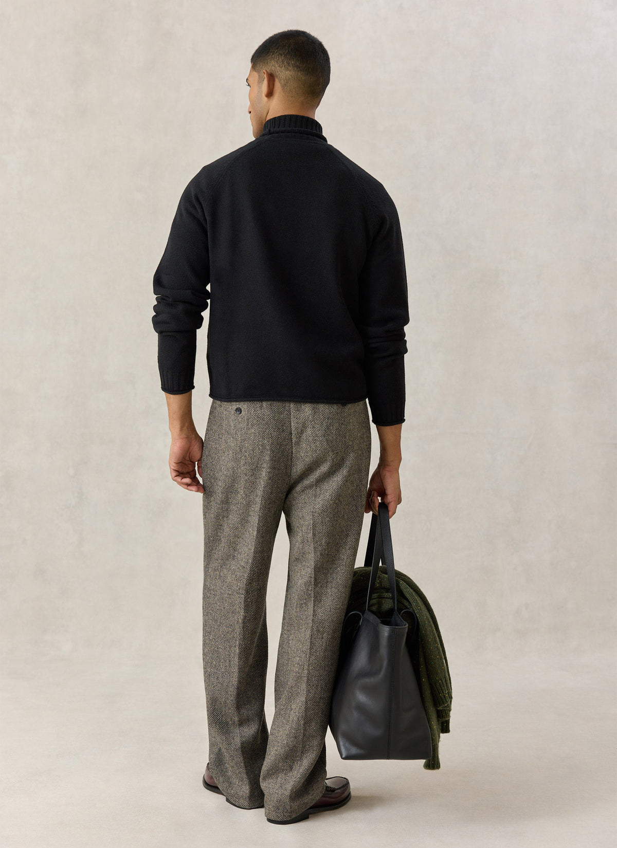 A model from the back wearing the Percival black roll-neck jumper, patterned trousers, and a black leather tote bag.