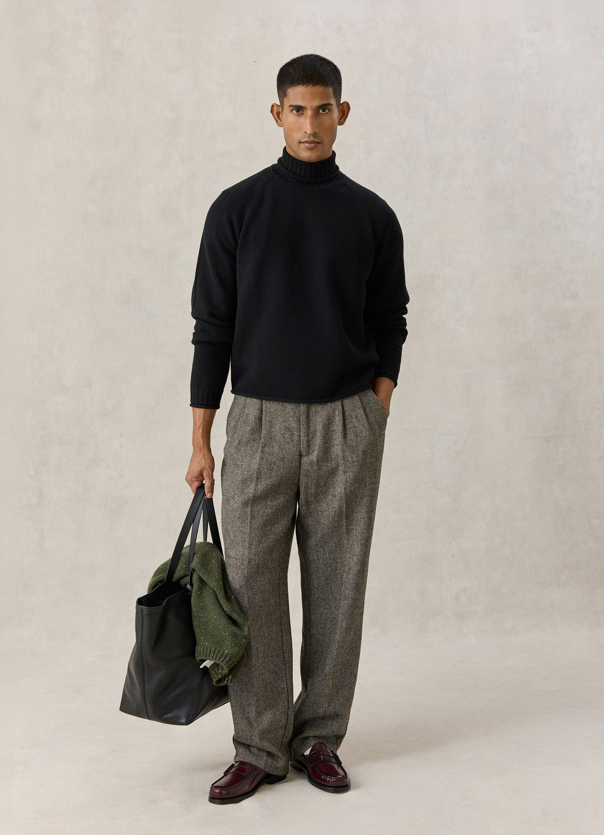 A model wearing the black Percival Dylan roll-neck jumper with trousers and loafers, holding a black leather tote bag.