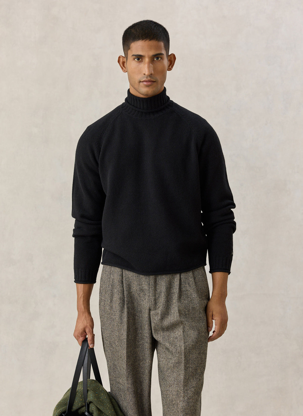 A model wearing the black Percival Dylan roll-neck jumper in 100% wool with patterned trousers and holding a bag.