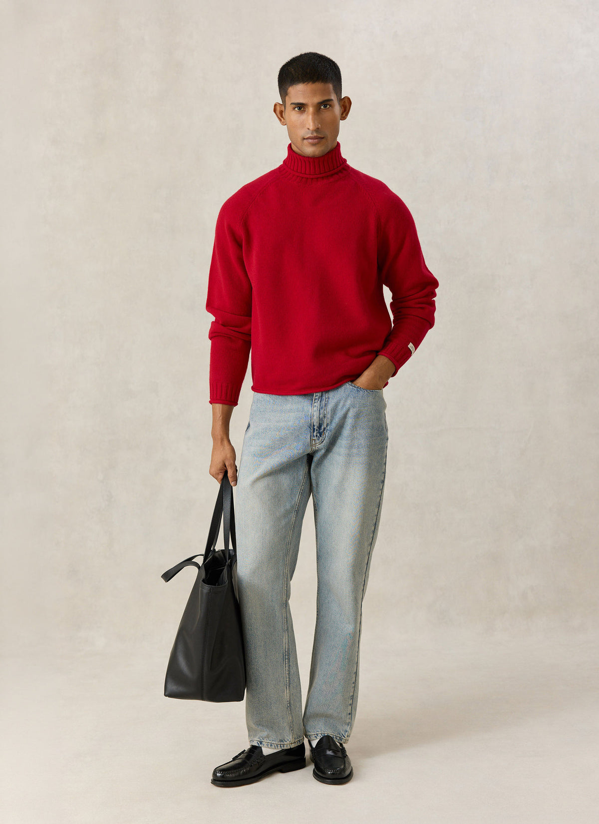 A model wearing the Percival red wool roll-neck jumper with light blue jeans and holding a black tote bag indoors.