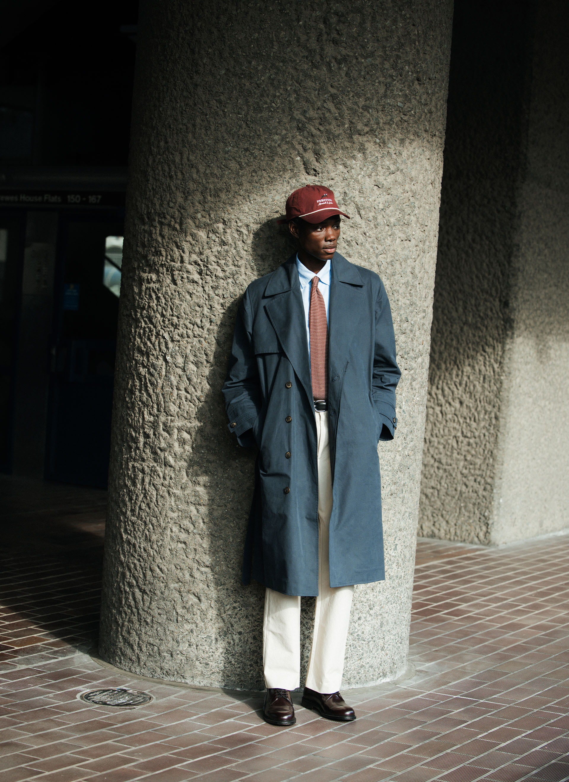 Navy cotton Percival x Front Office Mac Coat worn by a model leaning against a concrete pillar in an urban setting.