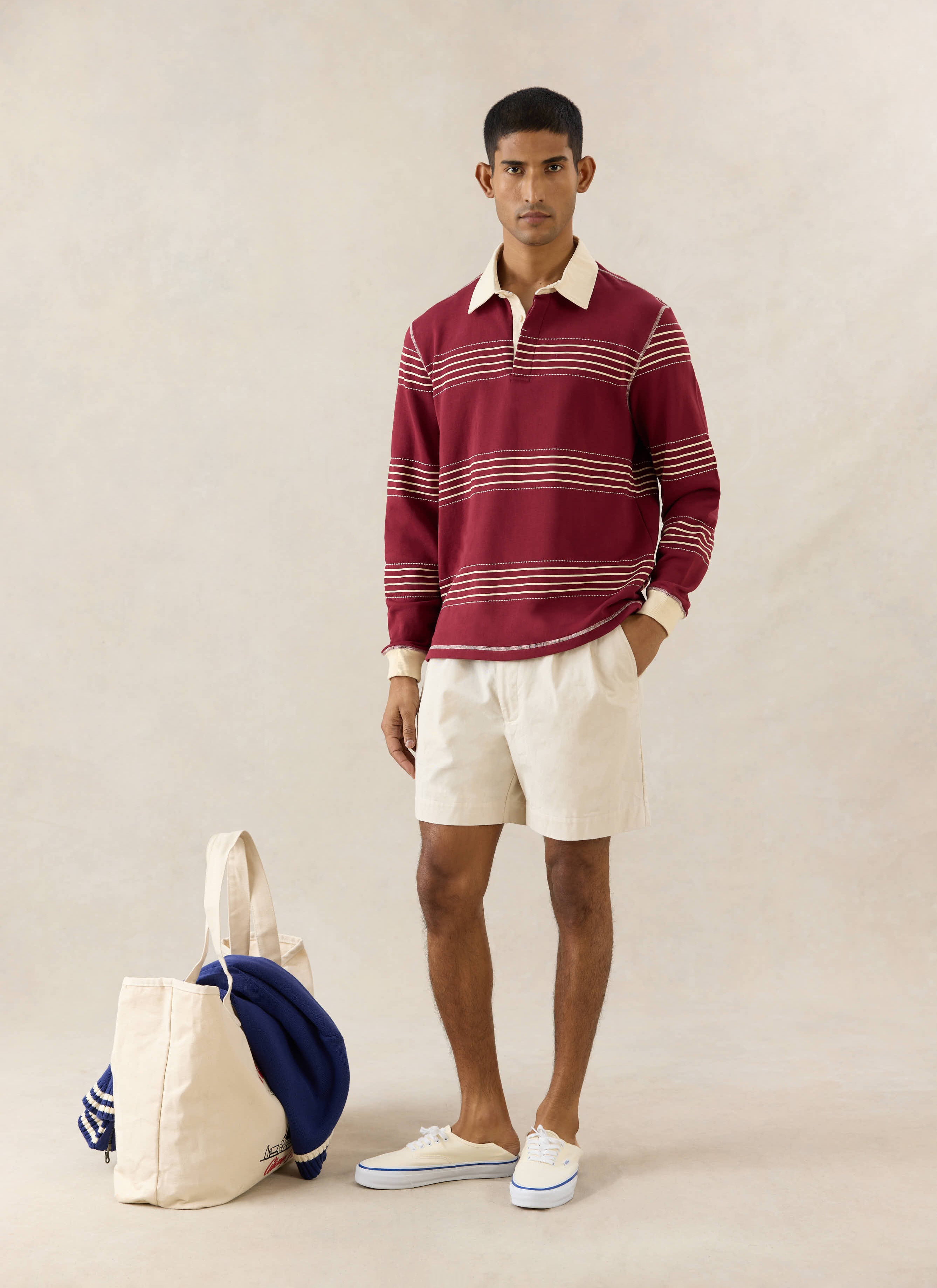 Percival Editor burgundy cotton rugby polo with cream stripes and stab stitching, styled with cream shorts and a tote bag.