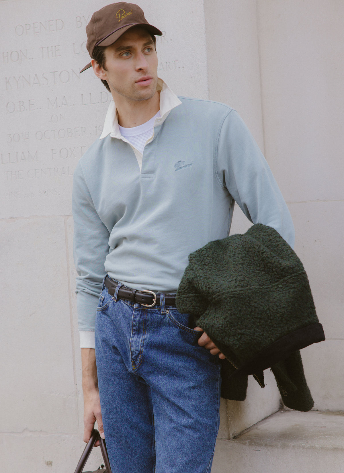 Model wearing the light blue Editor rugby polo with a cream collar, cap, jeans, and a dark fleece.