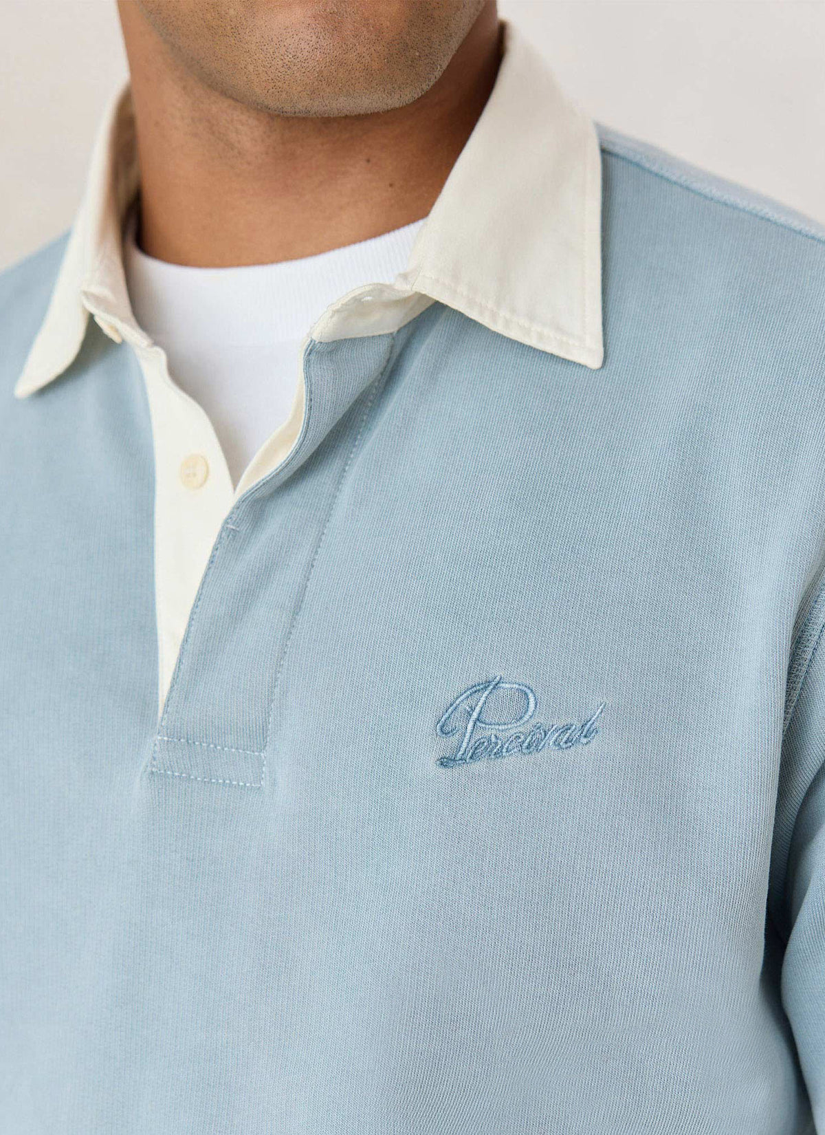 A close-up of the light blue Percival rugby polo shirt showing the contrasting cream collar and embroidered logo.