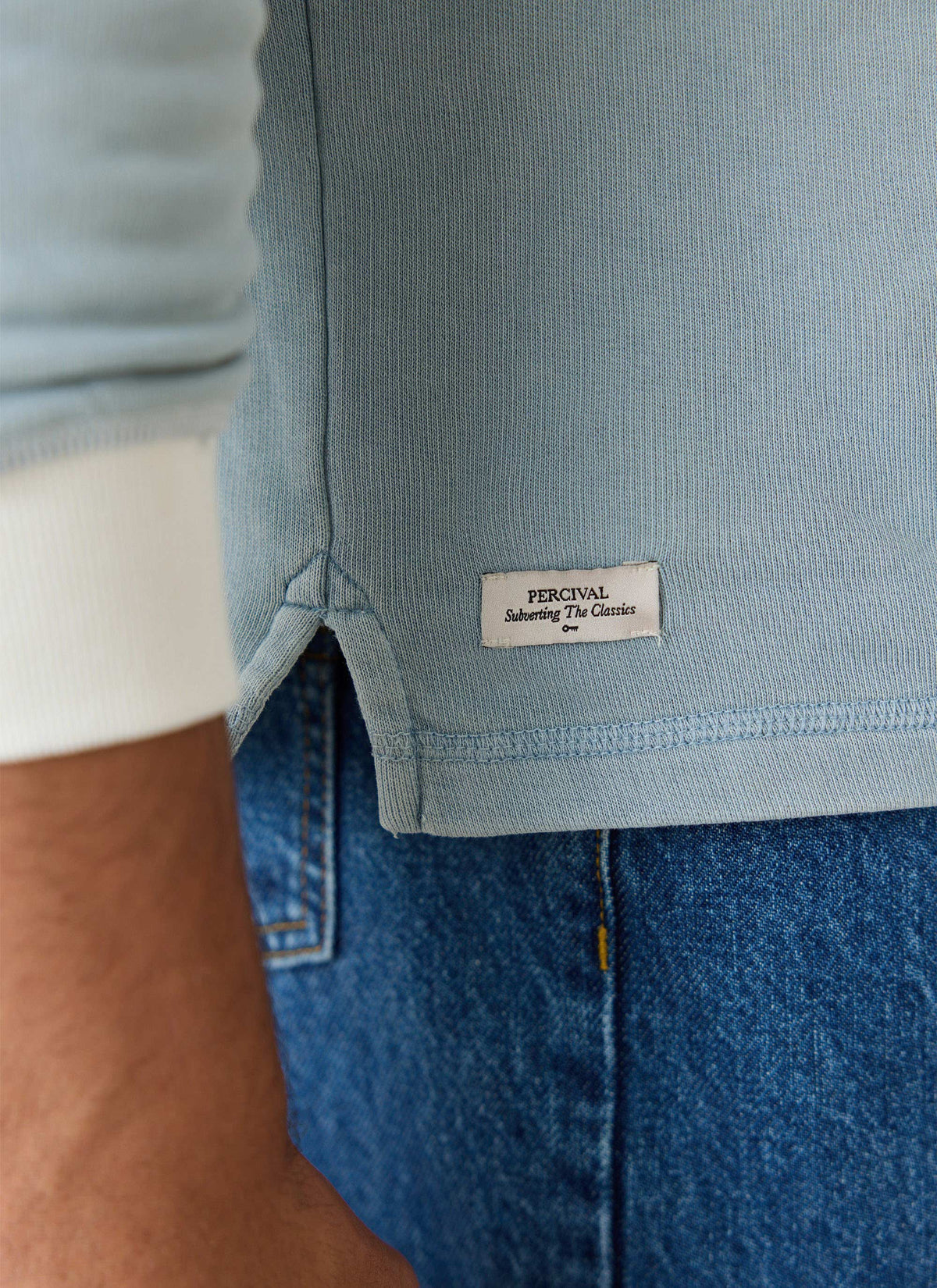 A close-up of the light blue Percival Editor rugby polo shirt, highlighting the woven hem tag and denim jeans.