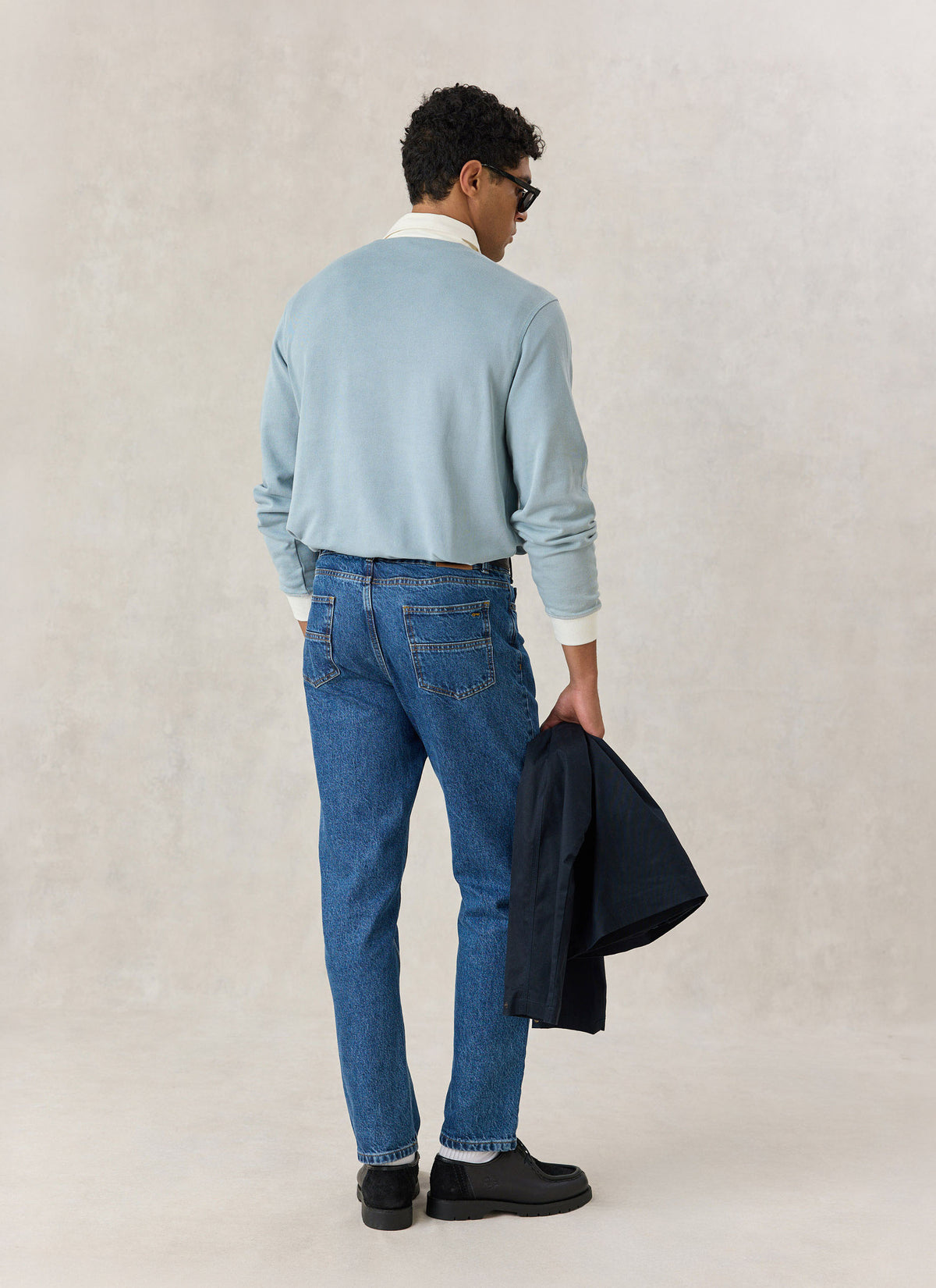 A model from behind wearing a light blue Percival rugby polo shirt and blue jeans, holding a dark jacket.