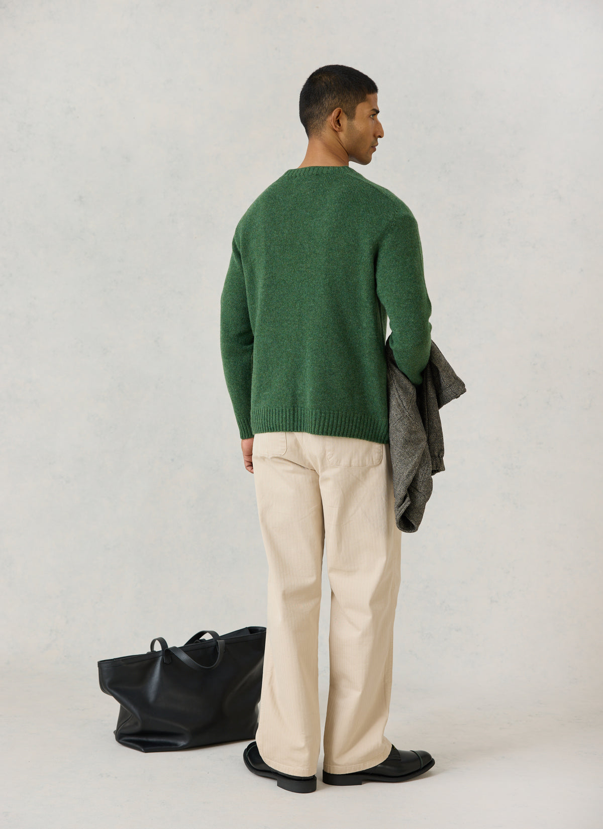 A model from the back wearing the Percival forest green lambswool jumper, cream trousers, and holding a coat.