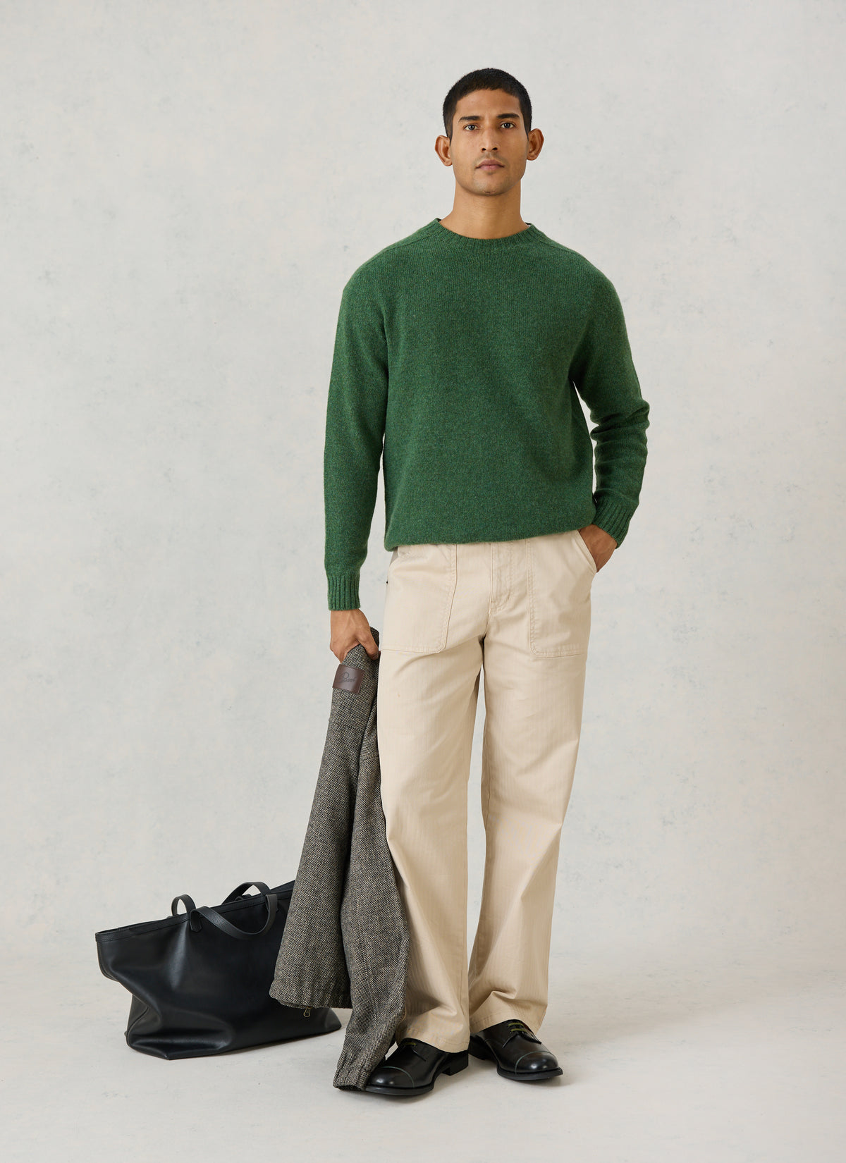 A model wearing the forest green Percival lambswool jumper with cream trousers, holding a jacket and a black leather bag.