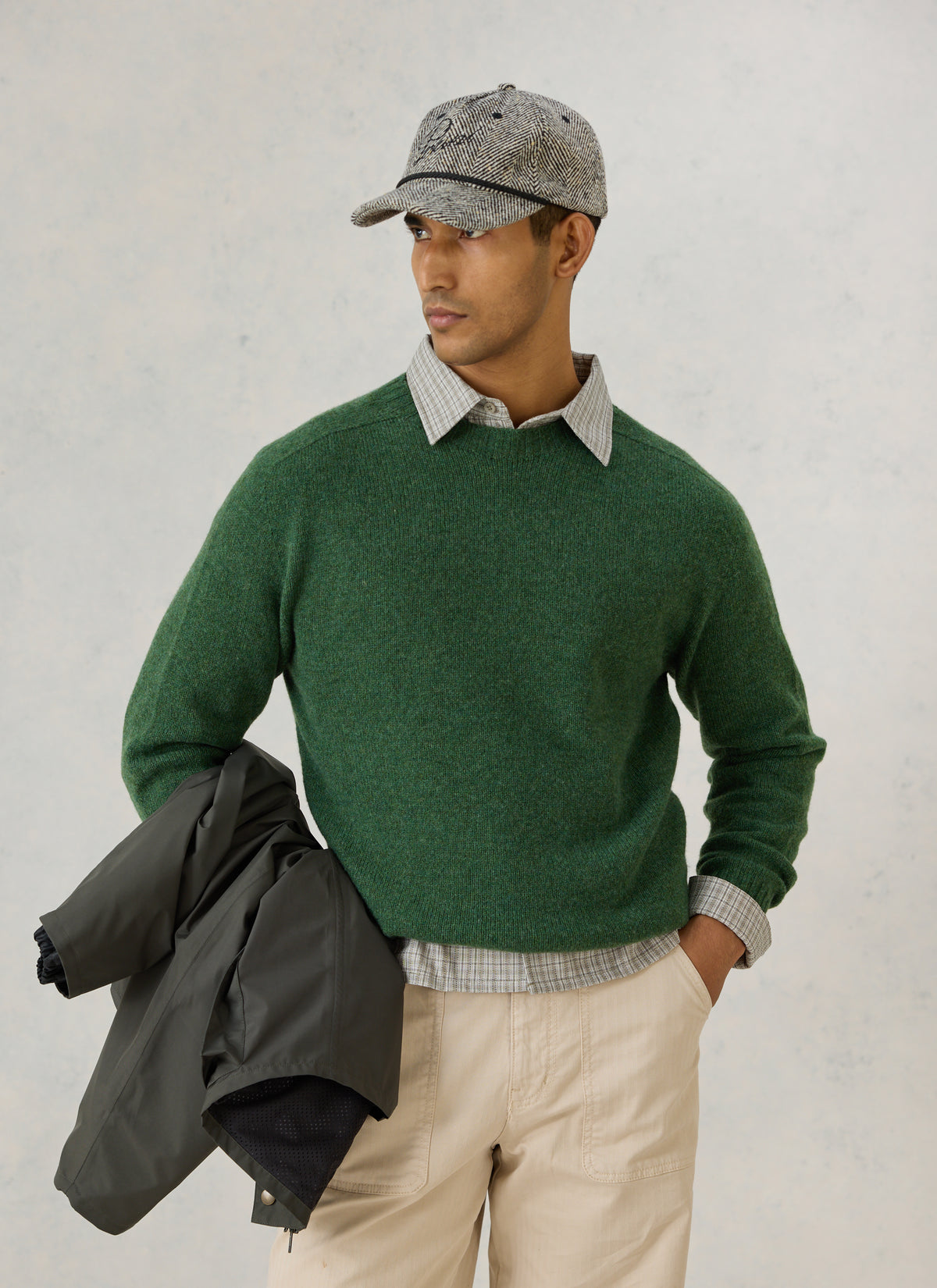 A model wearing the forest green lambswool jumper and grey herringbone cap by Percival, holding a dark jacket.
