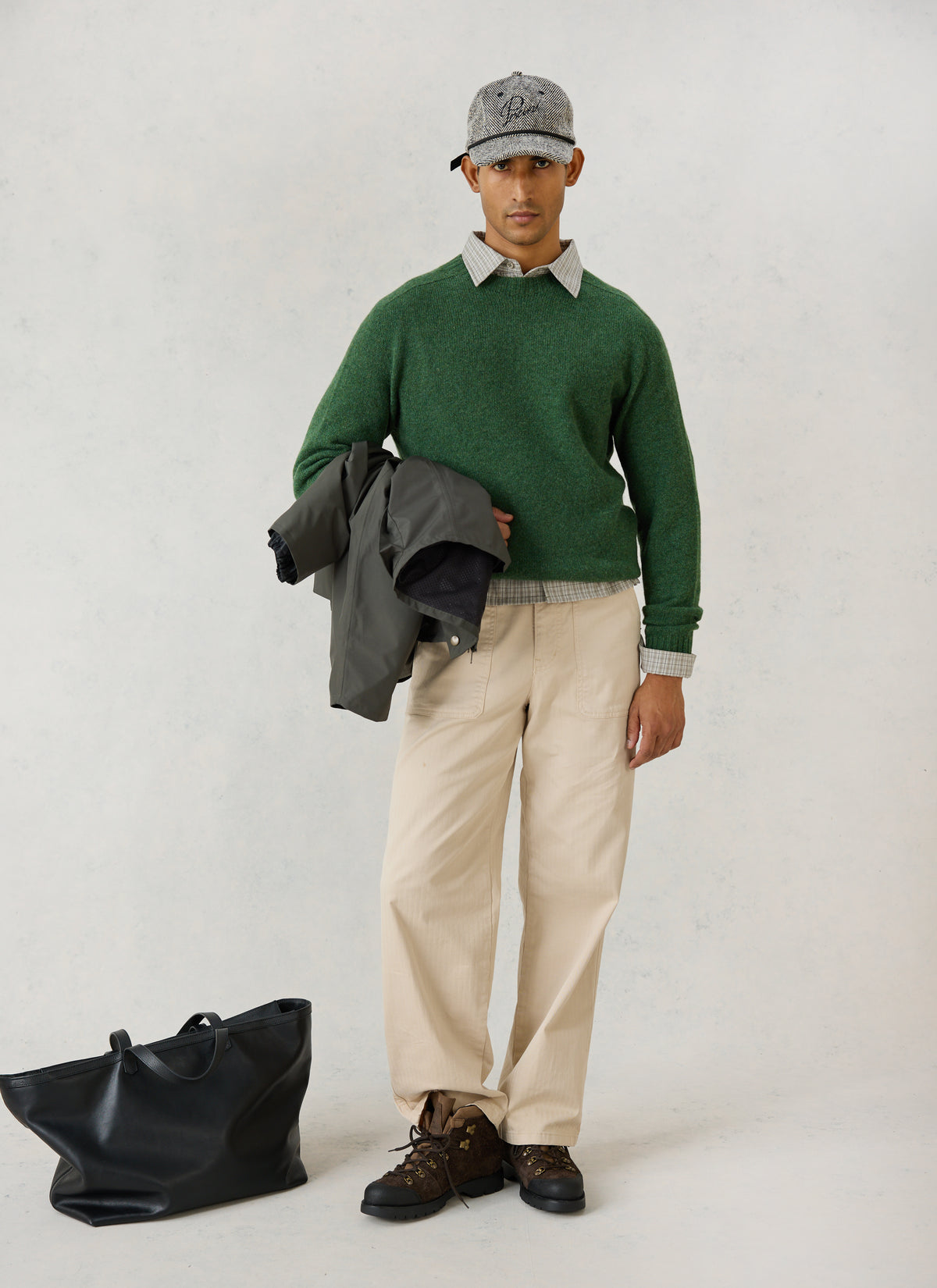 A model wearing the forest green lambswool jumper, grey herringbone trucker cap, cream trousers, and holding a dark jacket.