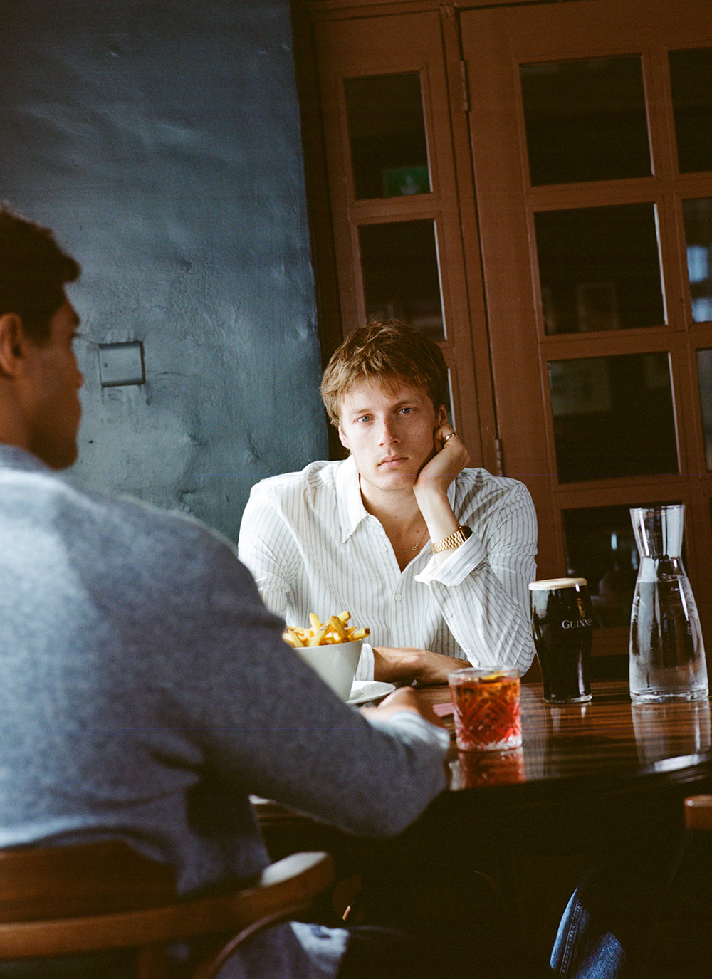 A model wears a white Percival striped shirt while sitting at a pub table with drinks.