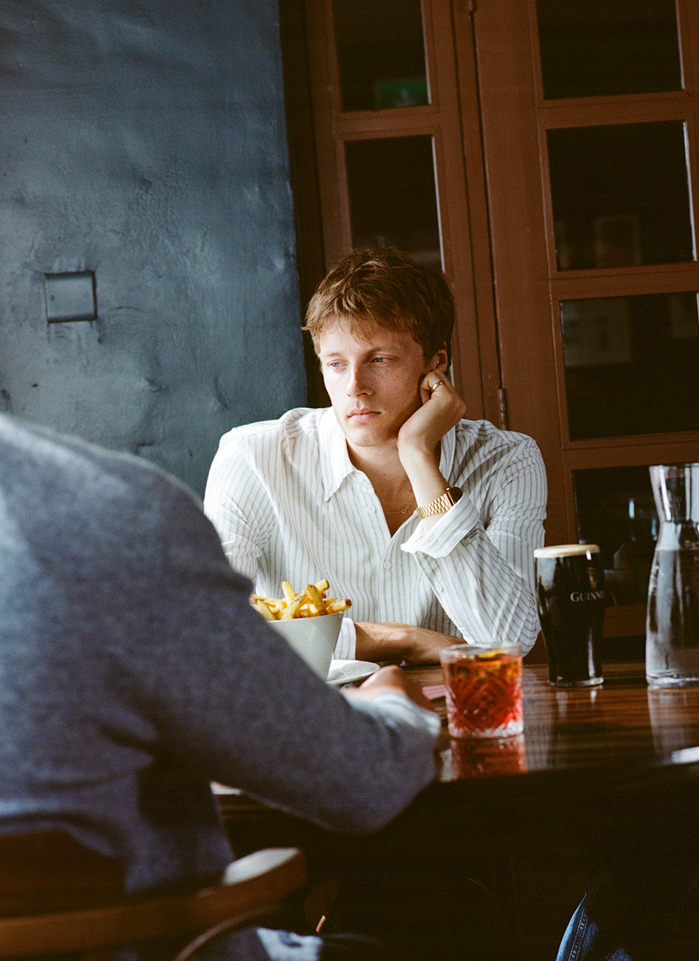 A model wears the white Fabian Striped Oxford Shirt from Percival, sitting at a pub table with drinks and chips.