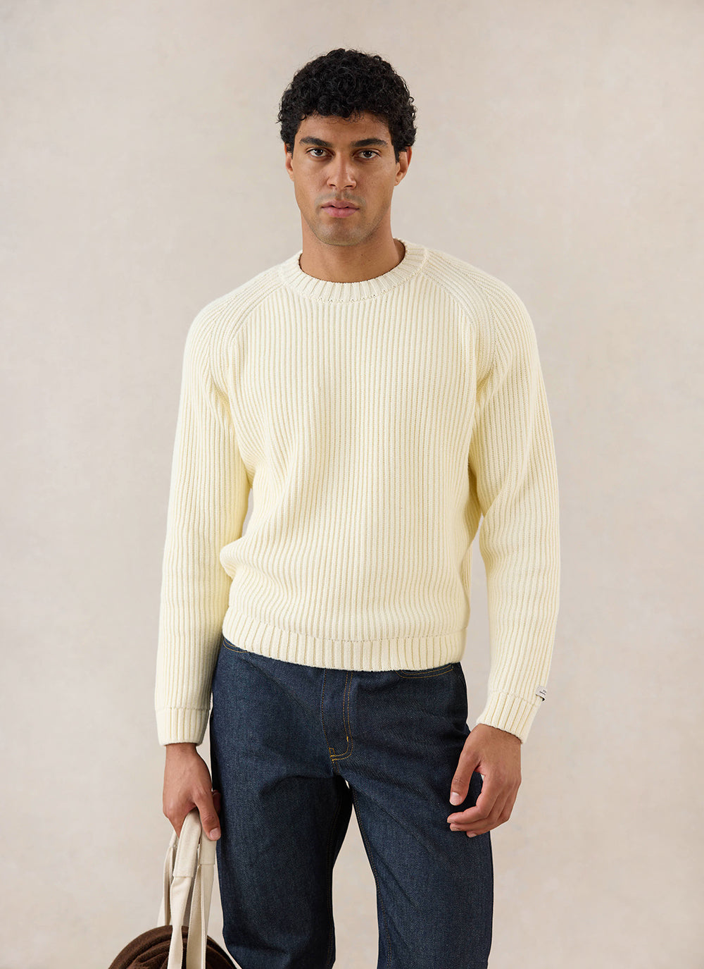 A model wears the cream Percival Fisherman Raglan Jumper, styled with dark denim jeans in a studio shot.