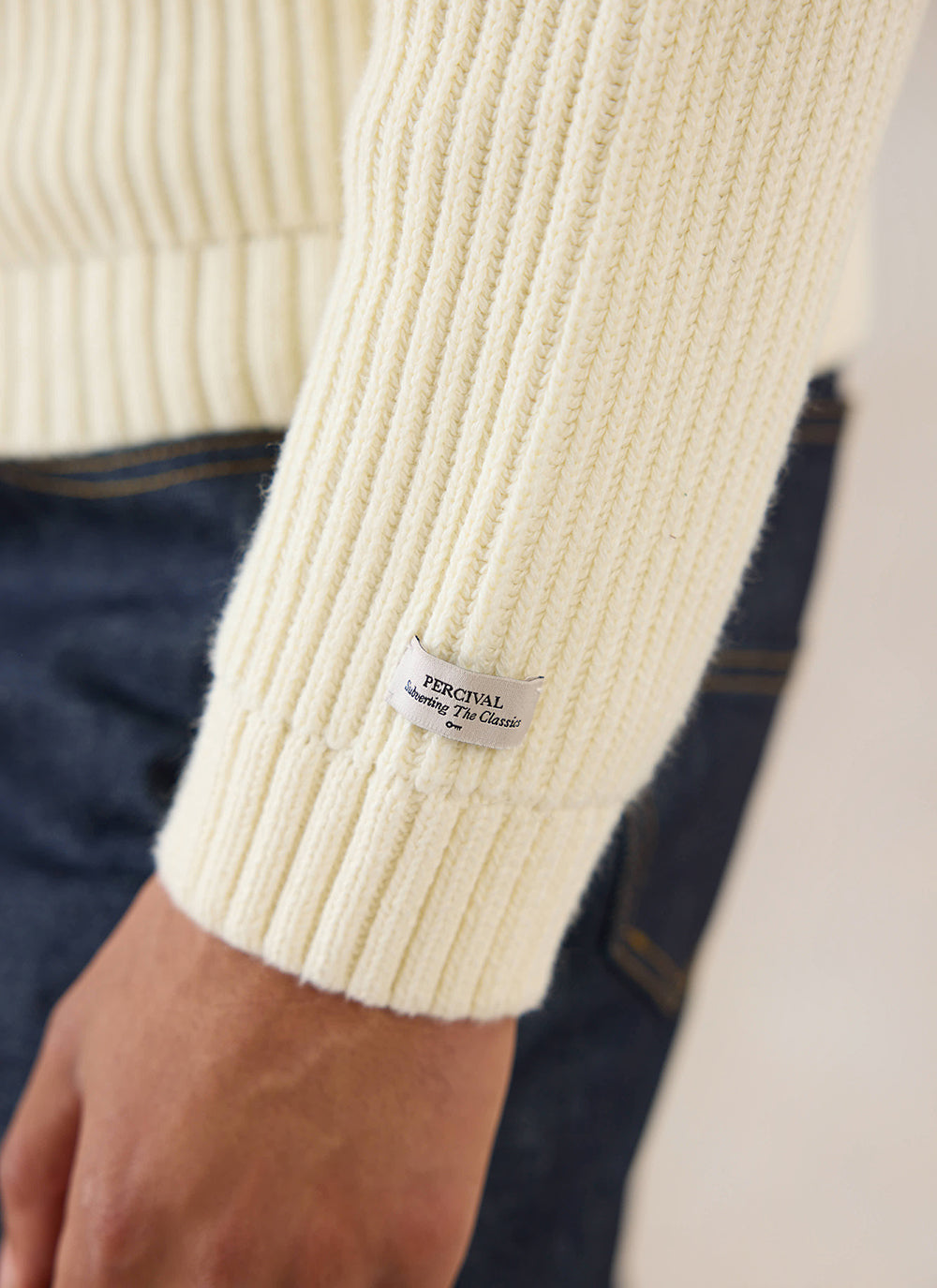 Close-up of the cream Percival Fisherman Knitted Jumper, highlighting the ribbed texture and branded sleeve label.
