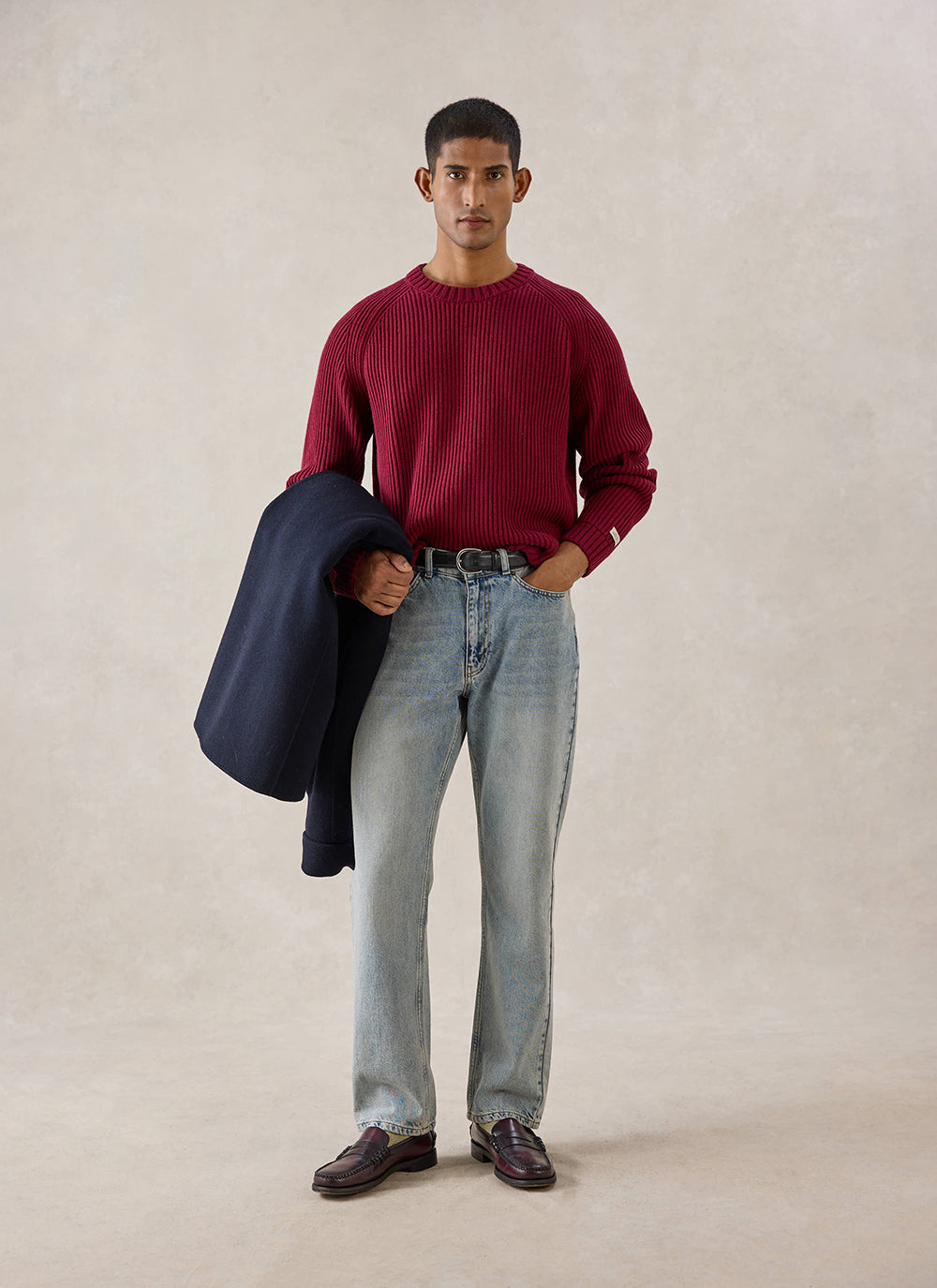 A model wears the merlot Percival Fisherman Knitted Jumper, styled with light wash blue jeans and dark loafers in a studio.