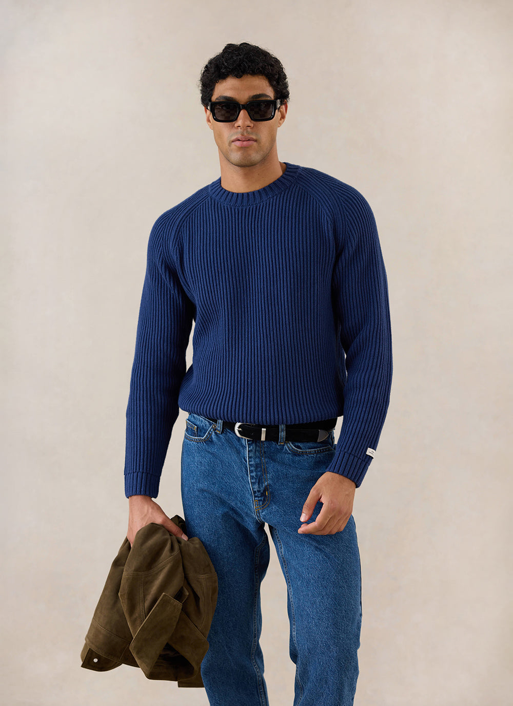 A model wears the navy Percival Fisherman Raglan Jumper with blue jeans, holding an olive jacket in a studio.