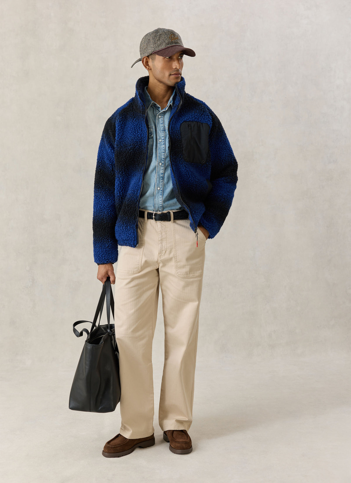 A model wearing the Percival ombre blue fleece jacket with a denim shirt, cream trousers, cap, and a black tote bag.