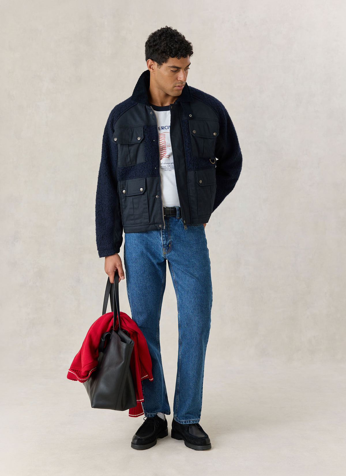 A model wearing the navy blue Percival Fletcher fleece and wax utility jacket with blue jeans and holding a dark tote bag.