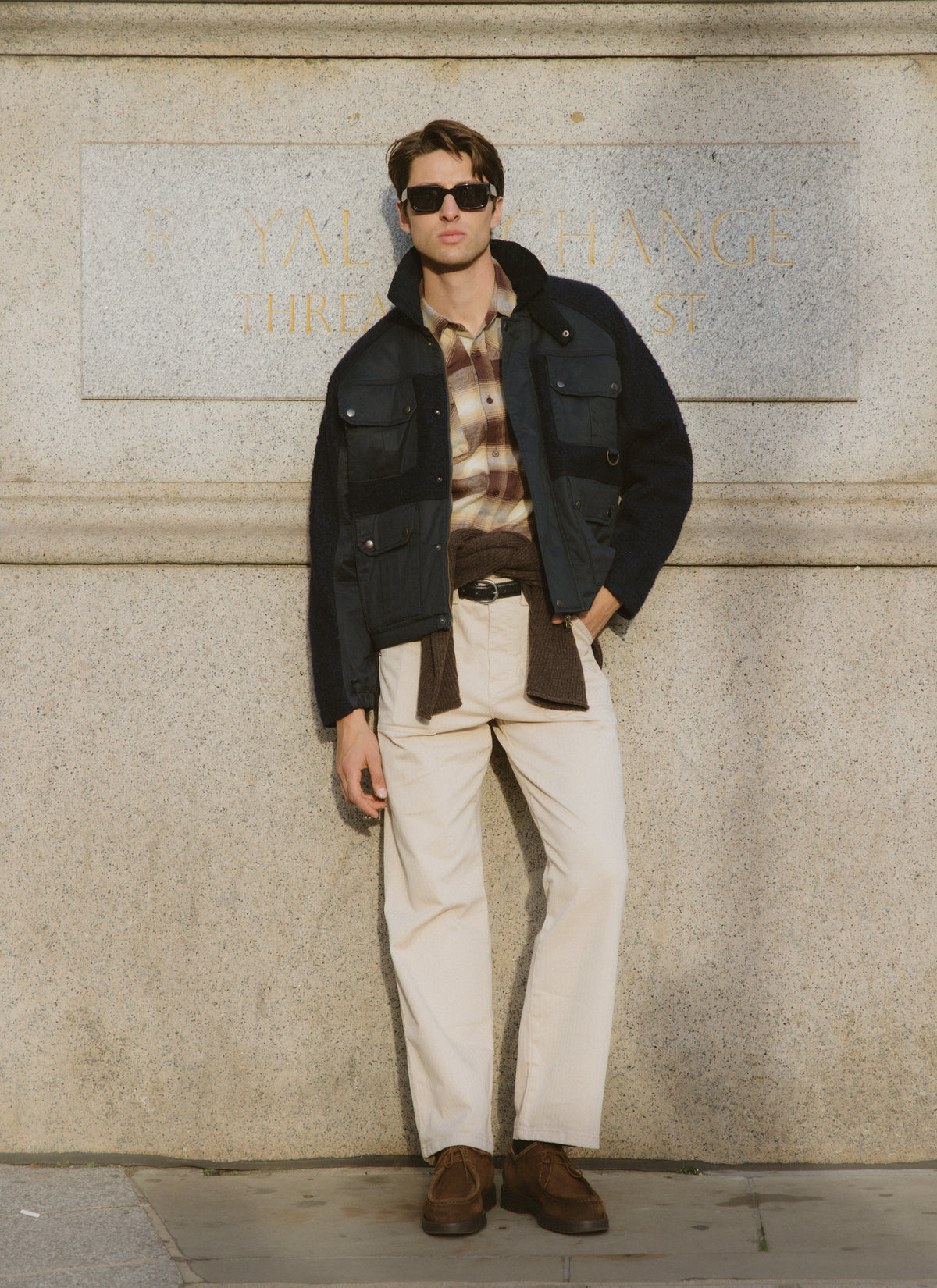 A model wearing a navy utility jacket over a brown checked western shirt while leaning against an outdoor stone wall.