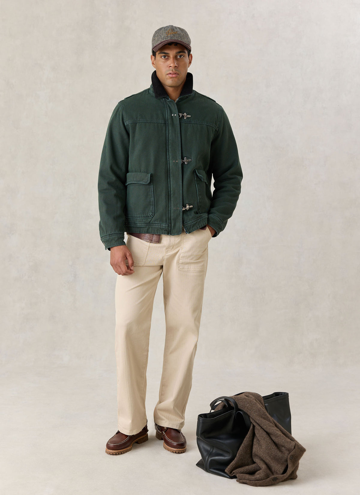 A model wearing the forest green Percival Fontane cotton fireman clasp jacket with trousers and a cap in a studio setting.