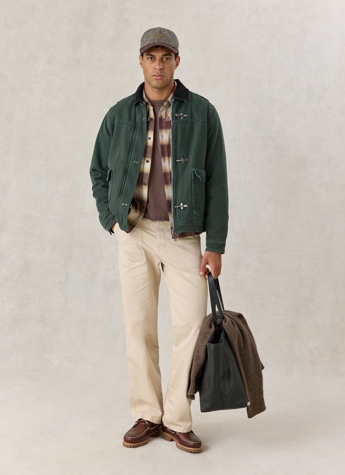 A model wearing the forest green Percival cotton fireman clasp jacket over a plaid shirt, beige trousers, and a cap.