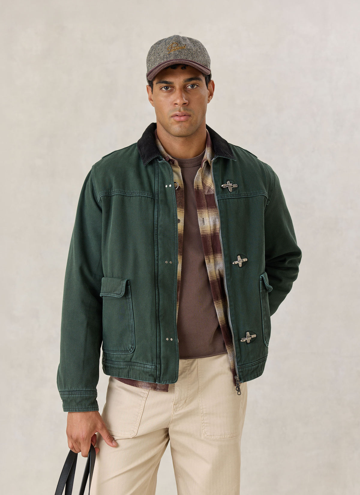 A model wearing the Percival forest green cotton fireman clasp jacket over a plaid shirt, cap, and beige trousers.