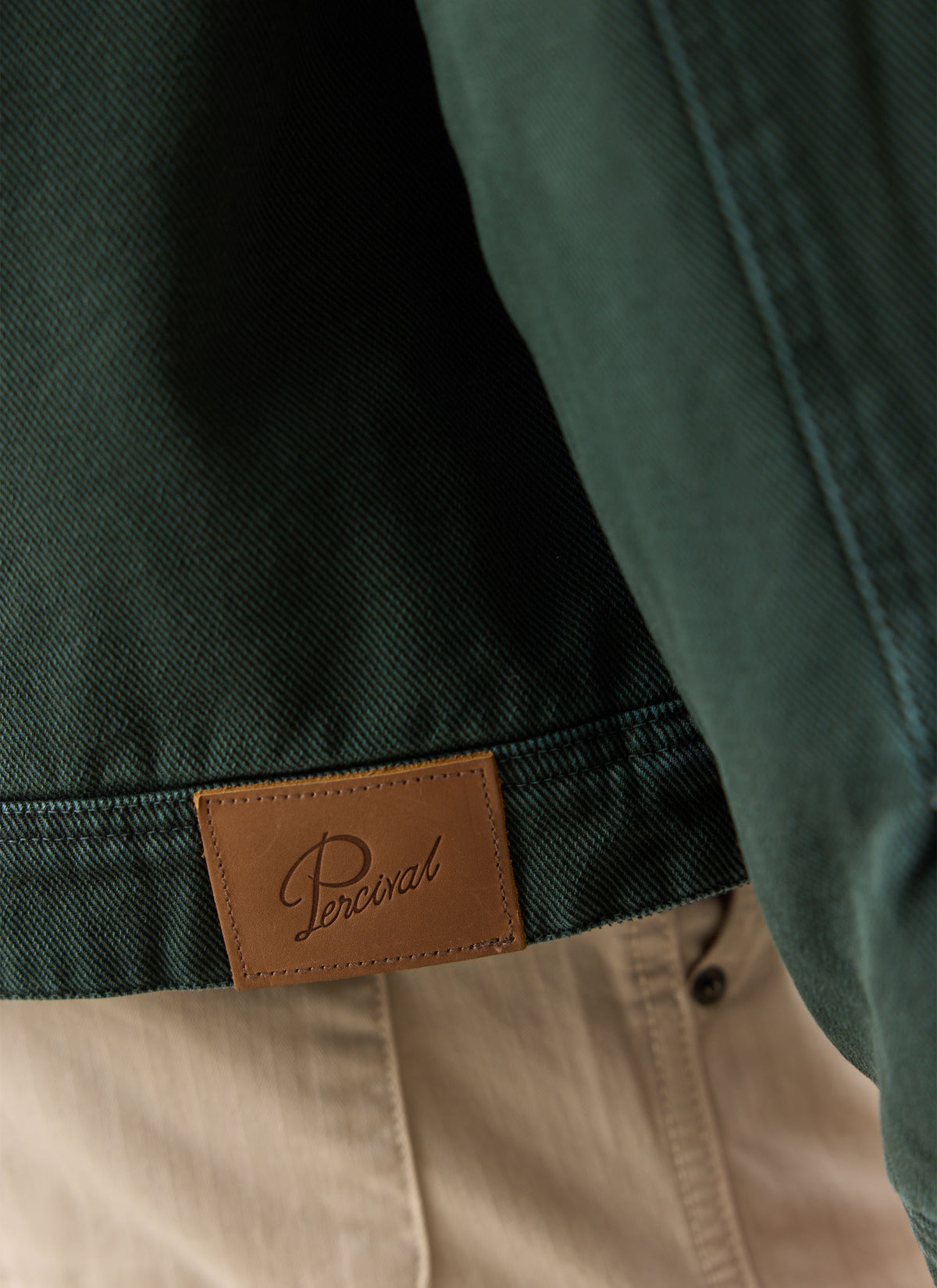A close-up of the forest green Percival cotton jacket showing the tan leather brand patch stitched on the hem.