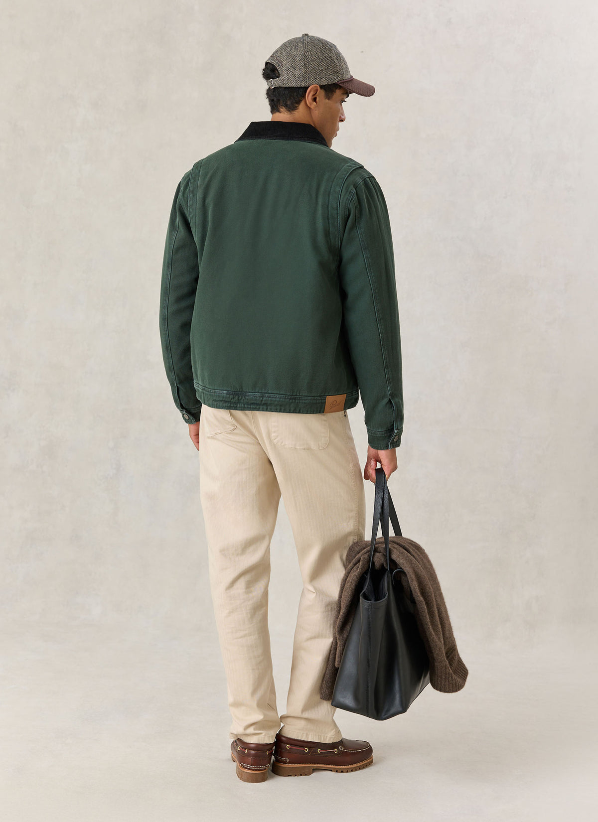 A model wearing the forest green Percival cotton jacket from behind, paired with light trousers and holding a black tote bag.