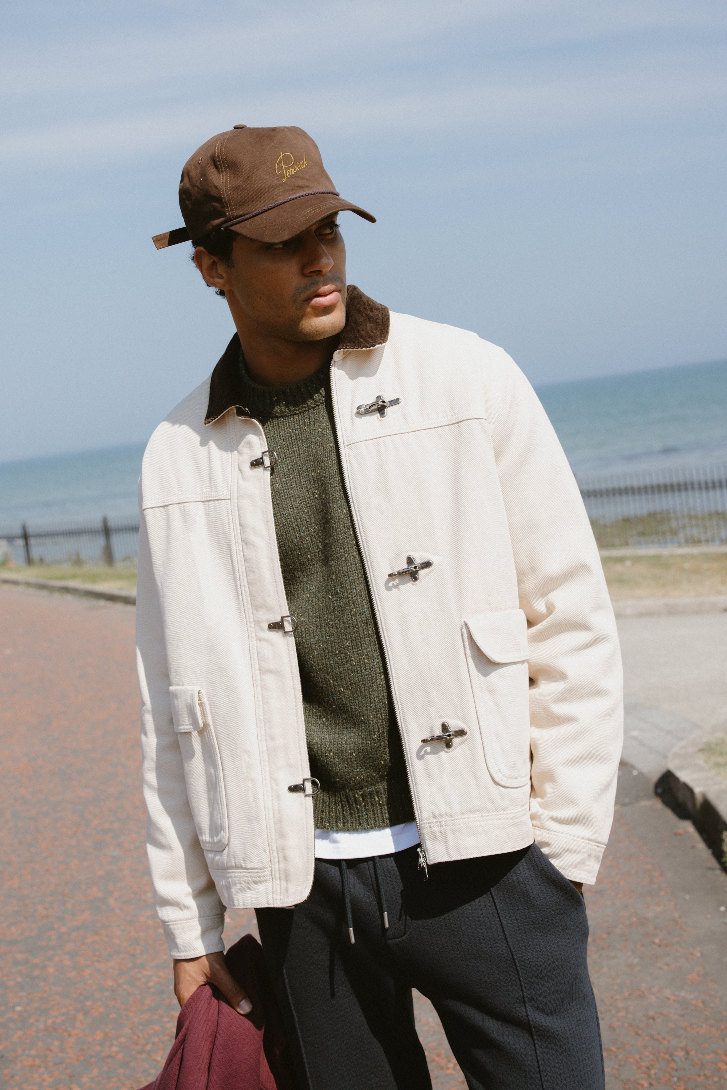 A model wears the cream Percival Fontane Fireman Clasp Jacket with metal hardware and corduroy collar by the sea.