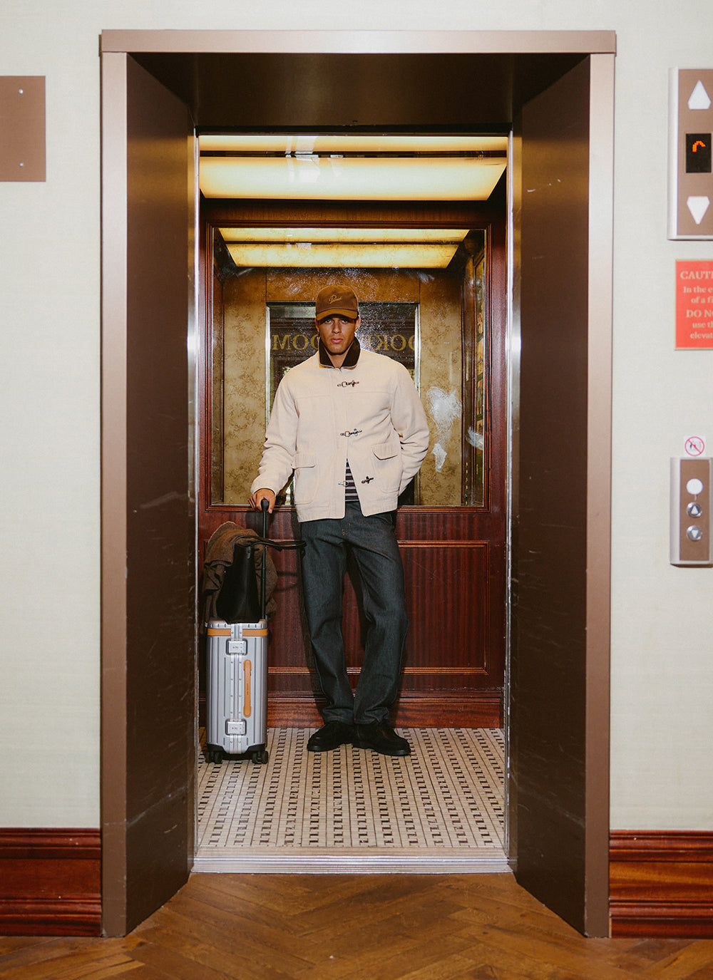 A model stands in an elevator wearing the cream Fontane jacket from Percival, paired with dark trousers and a cap.