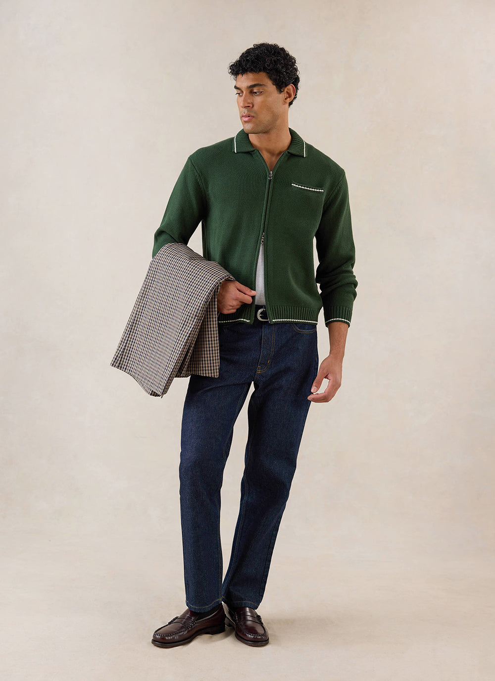Forest green Percival knitted zip cardigan in cotton, styled with dark denim jeans and a patterned jacket over the arm.