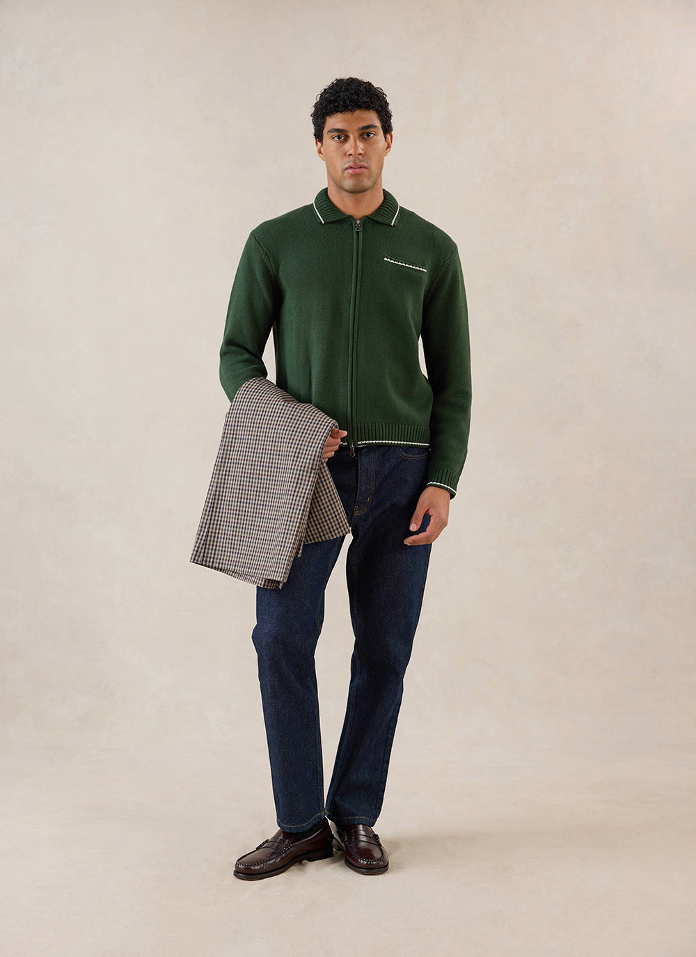 A model wearing the Percival forest green cotton zip cardigan, paired with dark jeans and holding a patterned jacket.