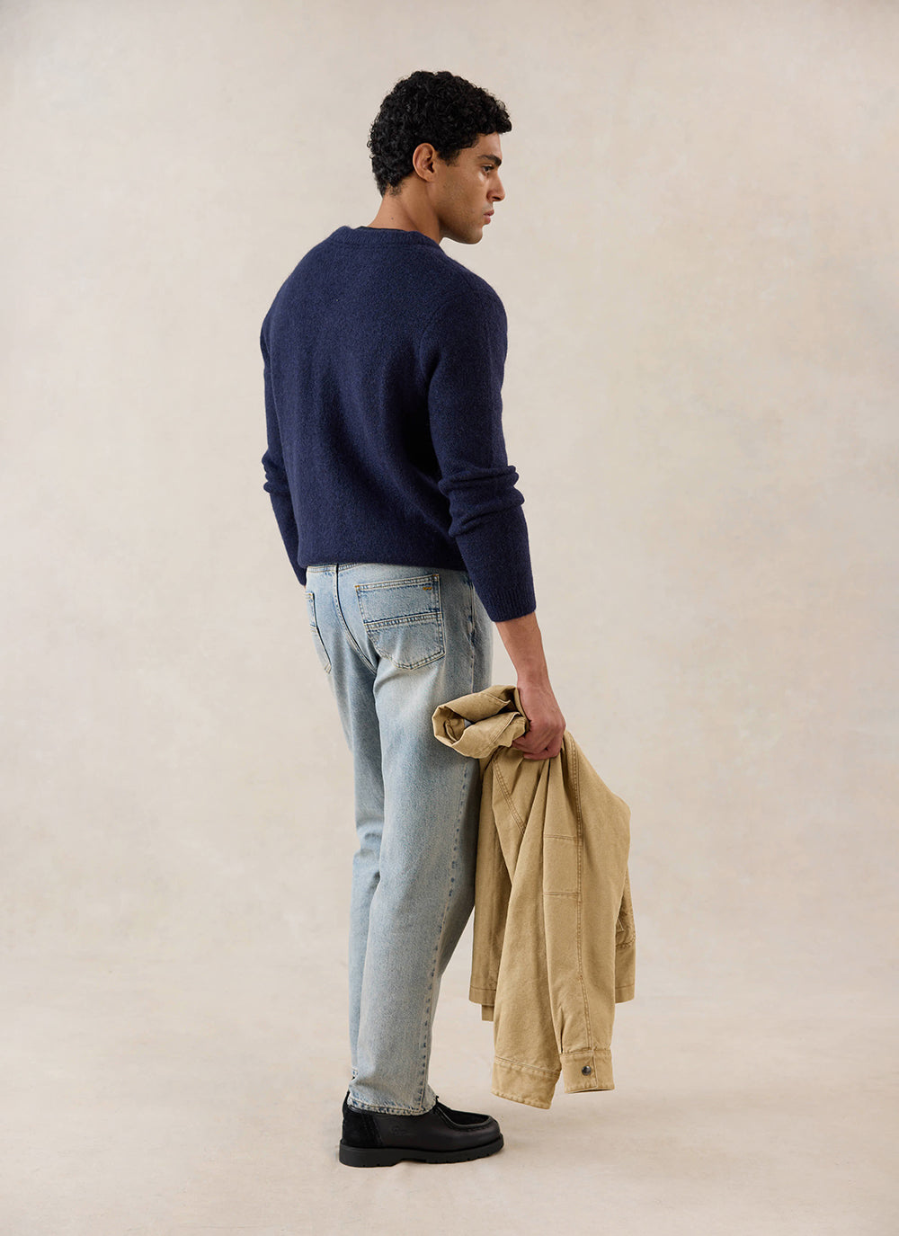 A model wears the navy Percival Gibson Cardigan from behind, with light wash jeans and carrying a tan jacket.