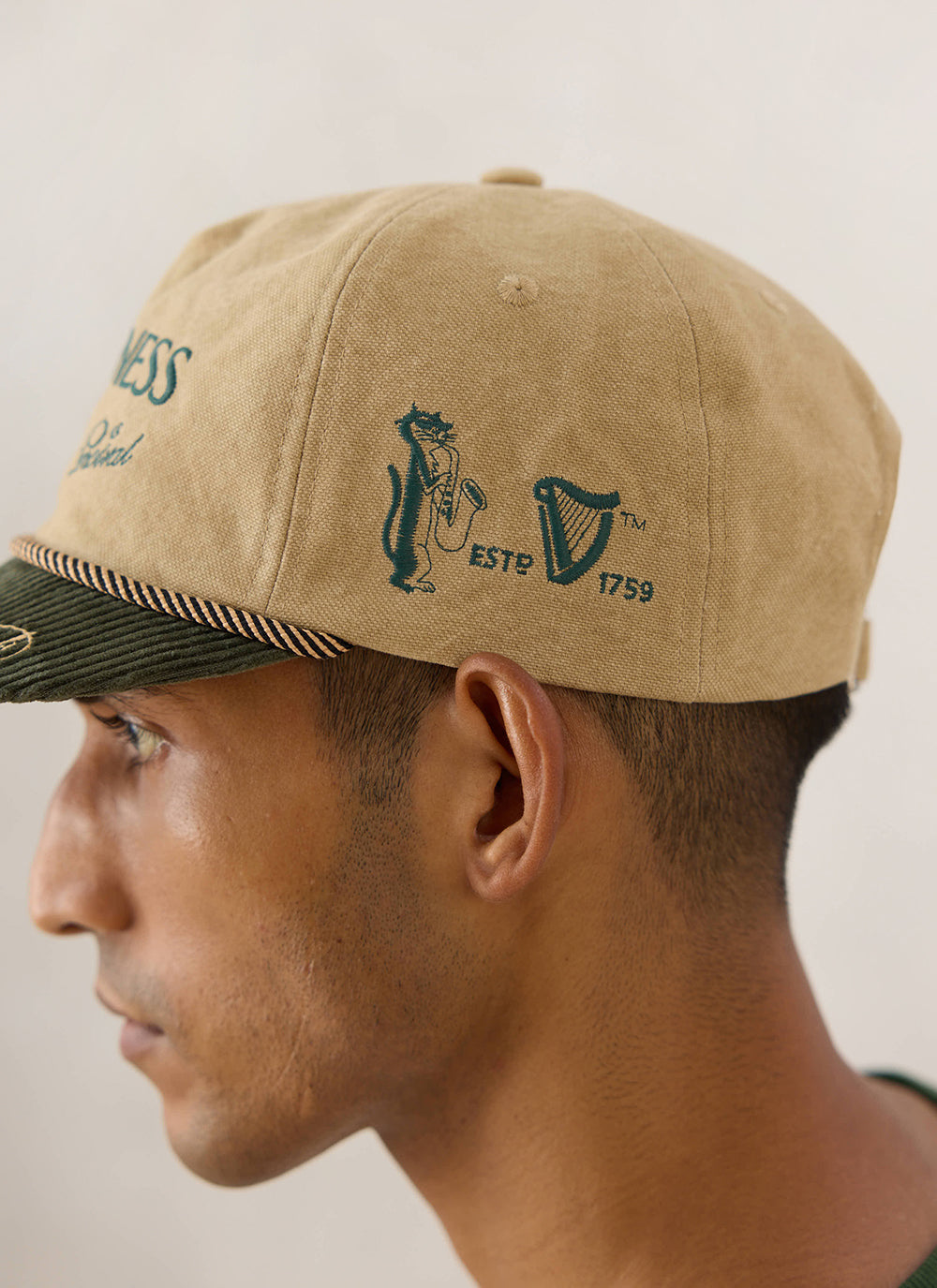 Side view of a model wearing the Guinness x Percival beige short peak cap, showing the green embroidered side graphics.