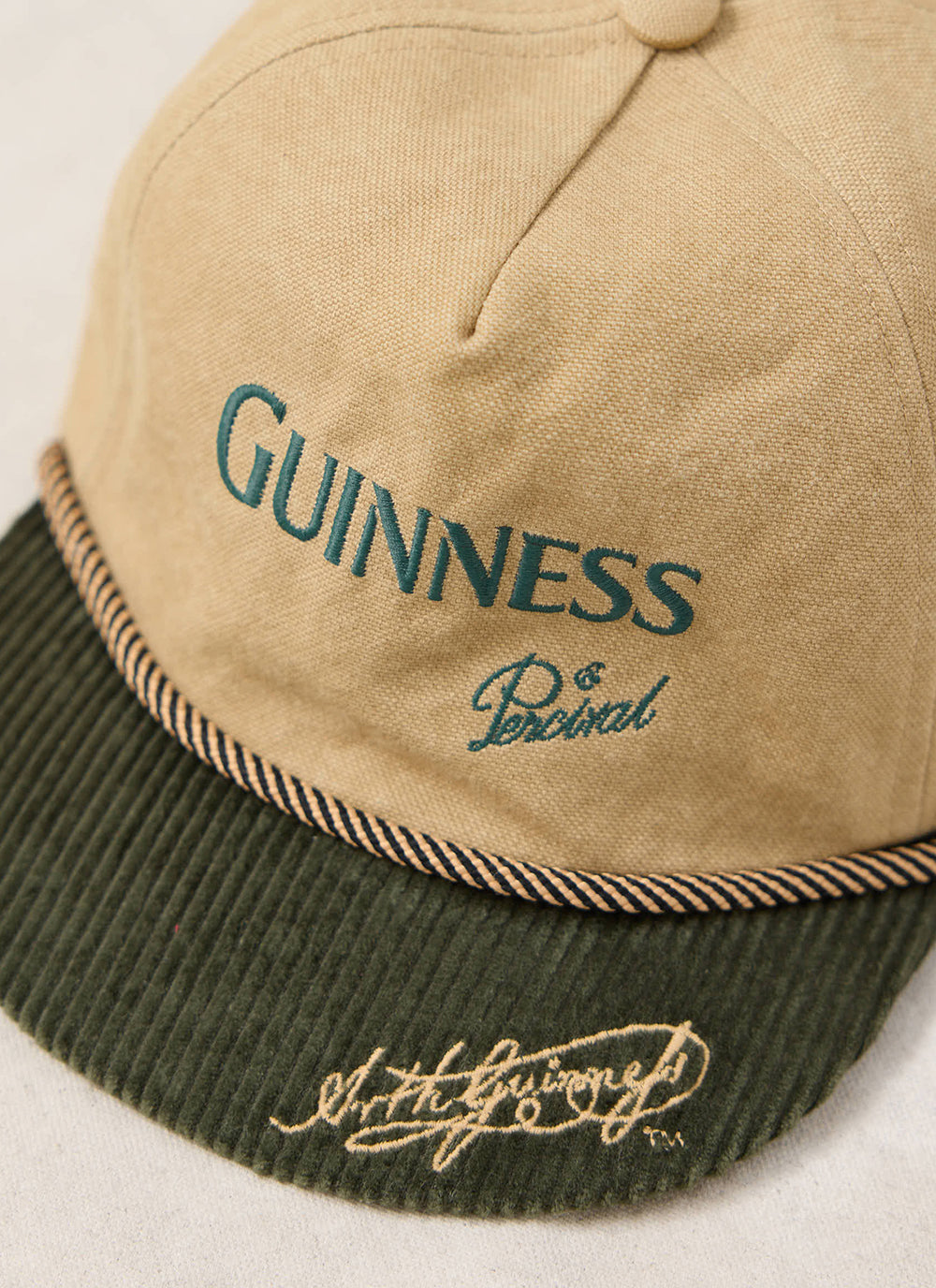 Close-up of the beige Guinness x Percival short peak cap, showing the green corduroy brim and front embroidery.