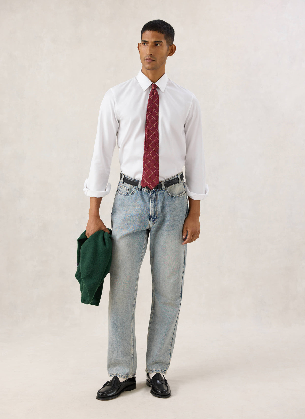 Model wearing the Guinness x Percival burgundy silk tie with a white shirt, blue jeans, and holding a green jumper.