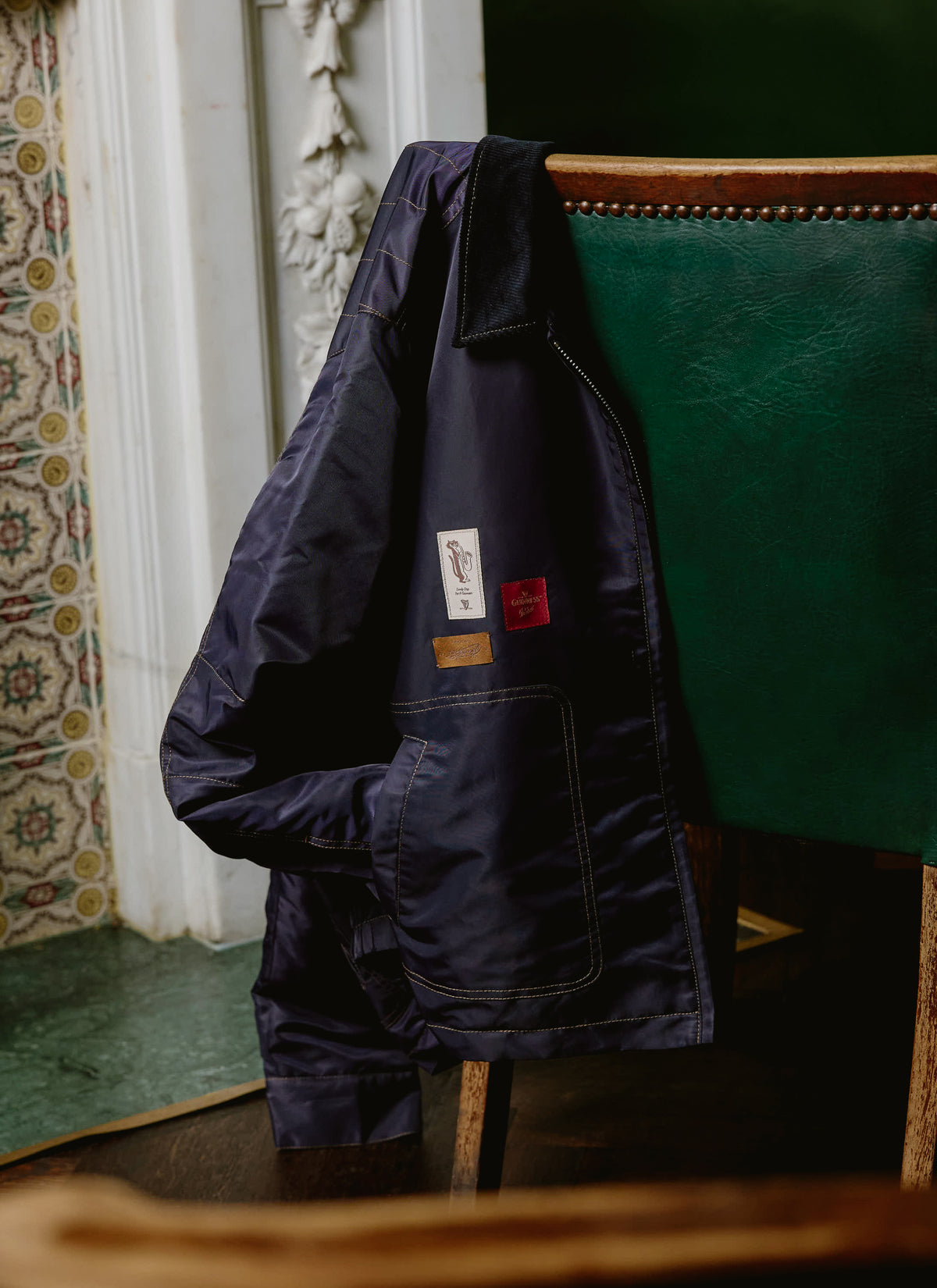 The Guinness x Percival navy jacket draped over a green chair, highlighting contrast stitching.