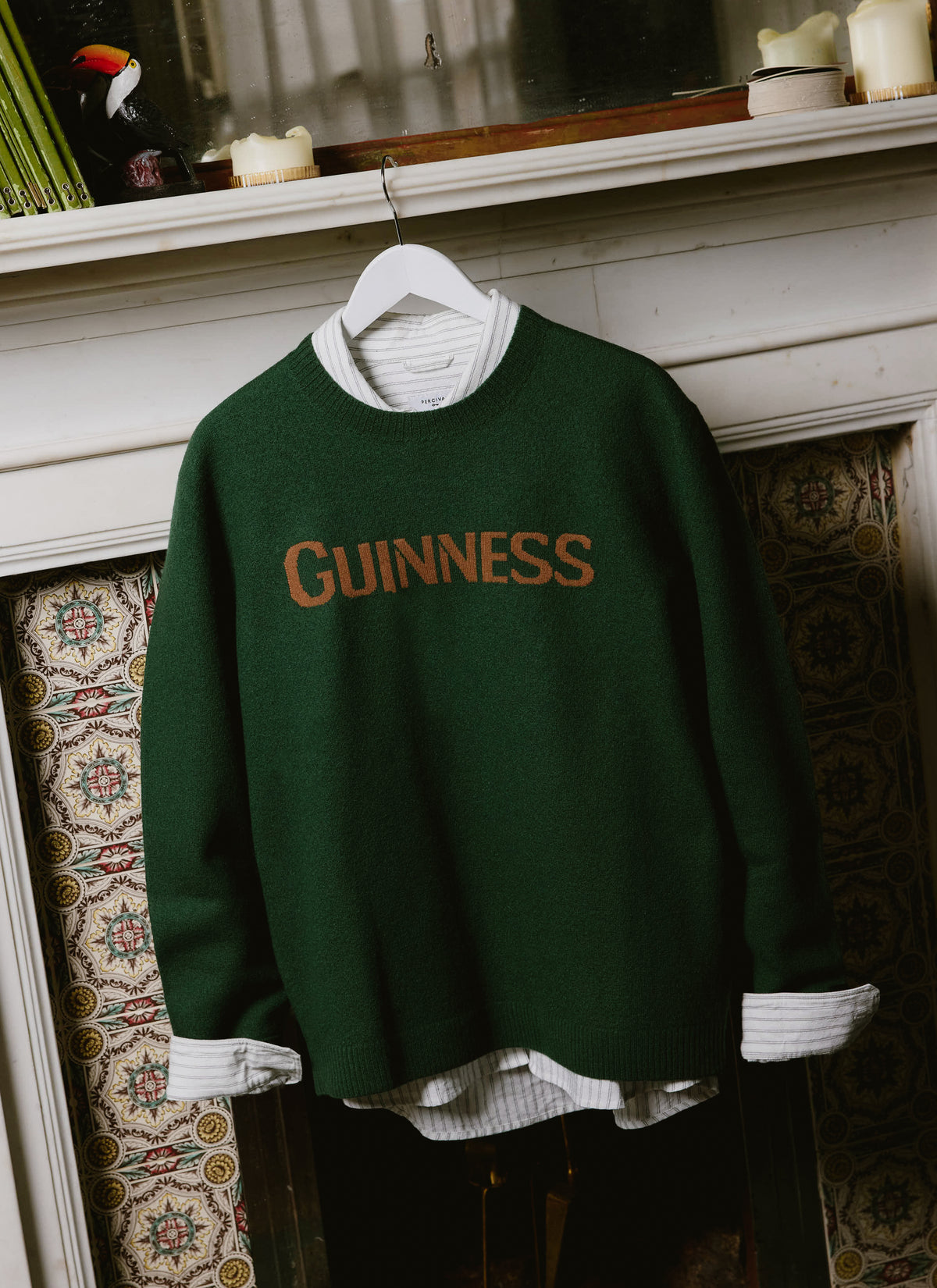 Guinness x Percival forest green merino wool jumper layered over a striped shirt, hanging against a decorative fireplace.