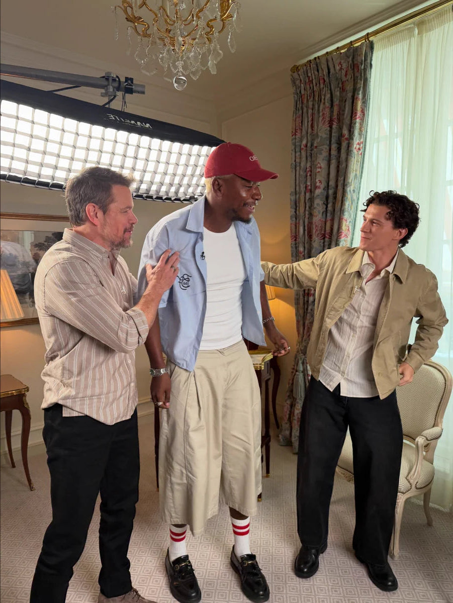 Three men posing together in an indoor room with curtains, a chandelier, and a studio light visible in the background. Tom Holland wears the Percival Lorenzo Shirt.