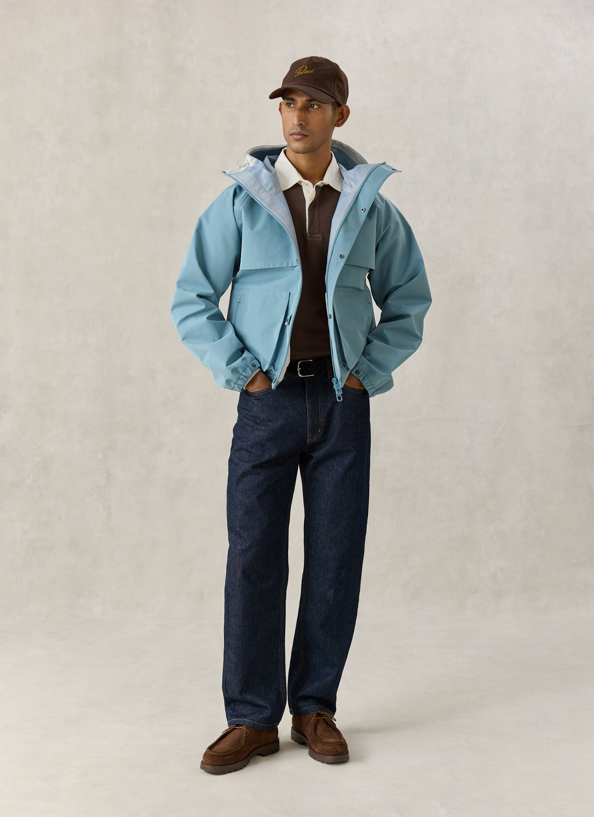 A model wearing the Percival light blue waterproof Hanson rain jacket in a boxy fit over a brown polo and dark jeans.