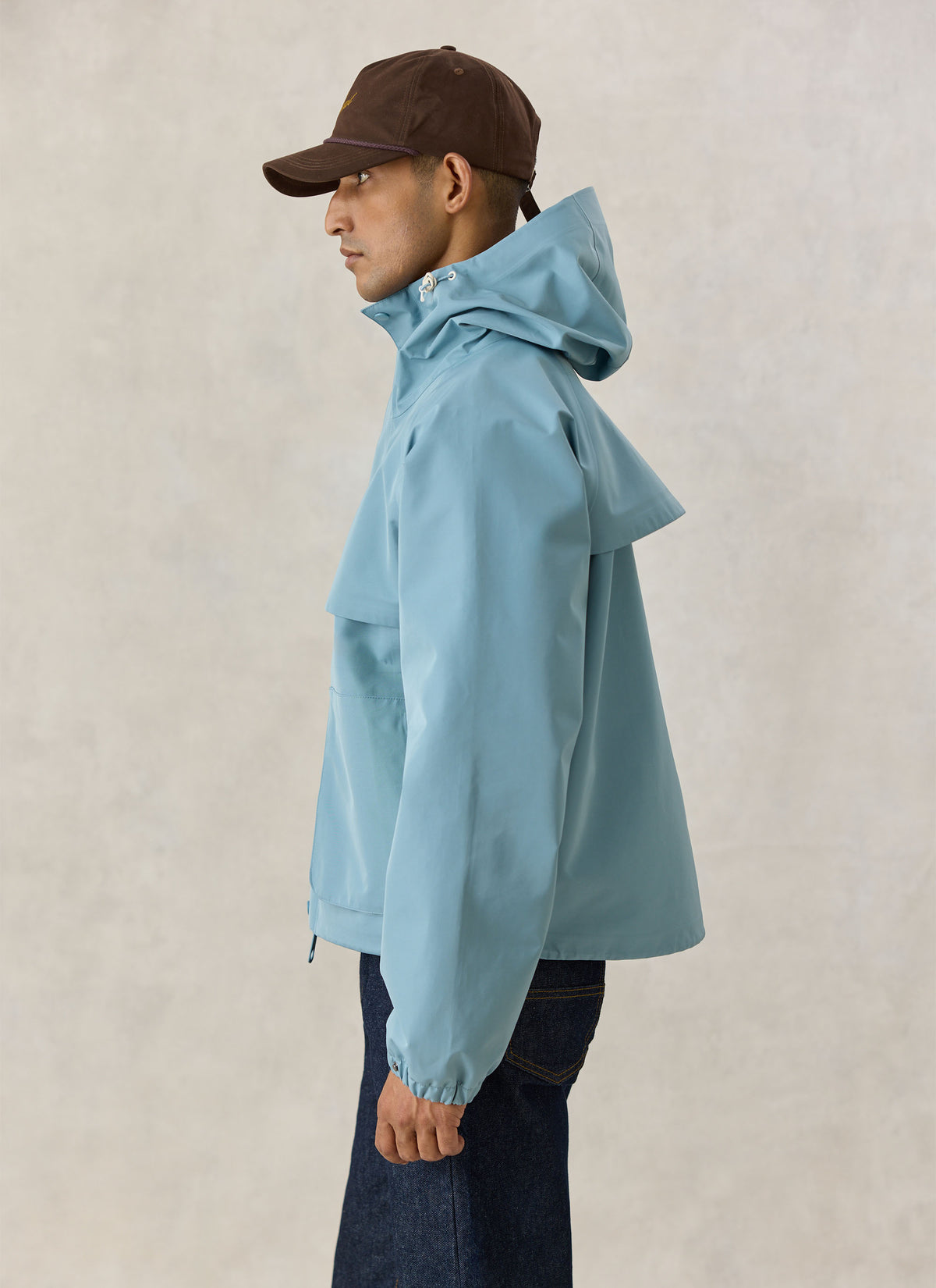 A model wearing the Percival Hanson boxy waterproof rain jacket in light blue with a cap.