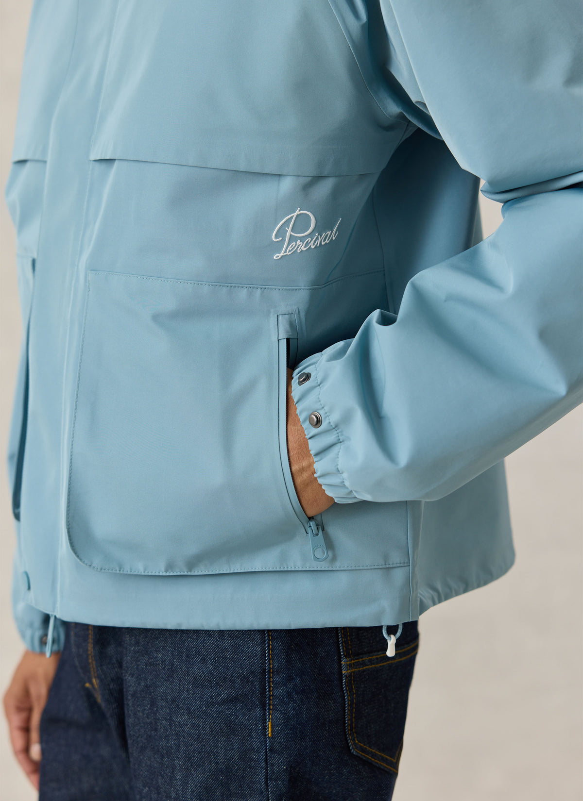 Close-up of the Percival Hanson waterproof rain jacket in light blue showing the chest pocket and embroidered logo.