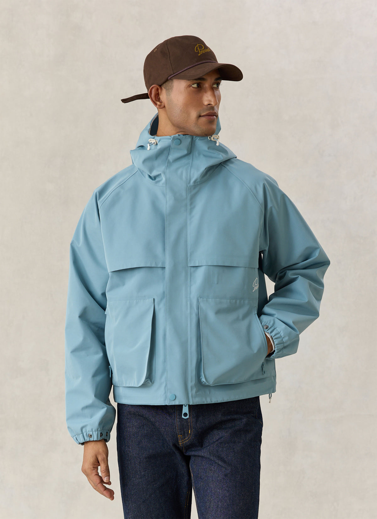 A model wearing the light blue Percival Hanson waterproof boxy rain jacket with a hood, large pockets, and a brown cap.