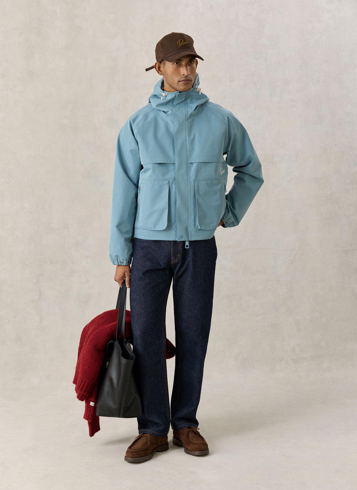 A model wearing the Percival light blue Hanson boxy waterproof rain jacket with dark jeans, a cap, and holding a tote bag.