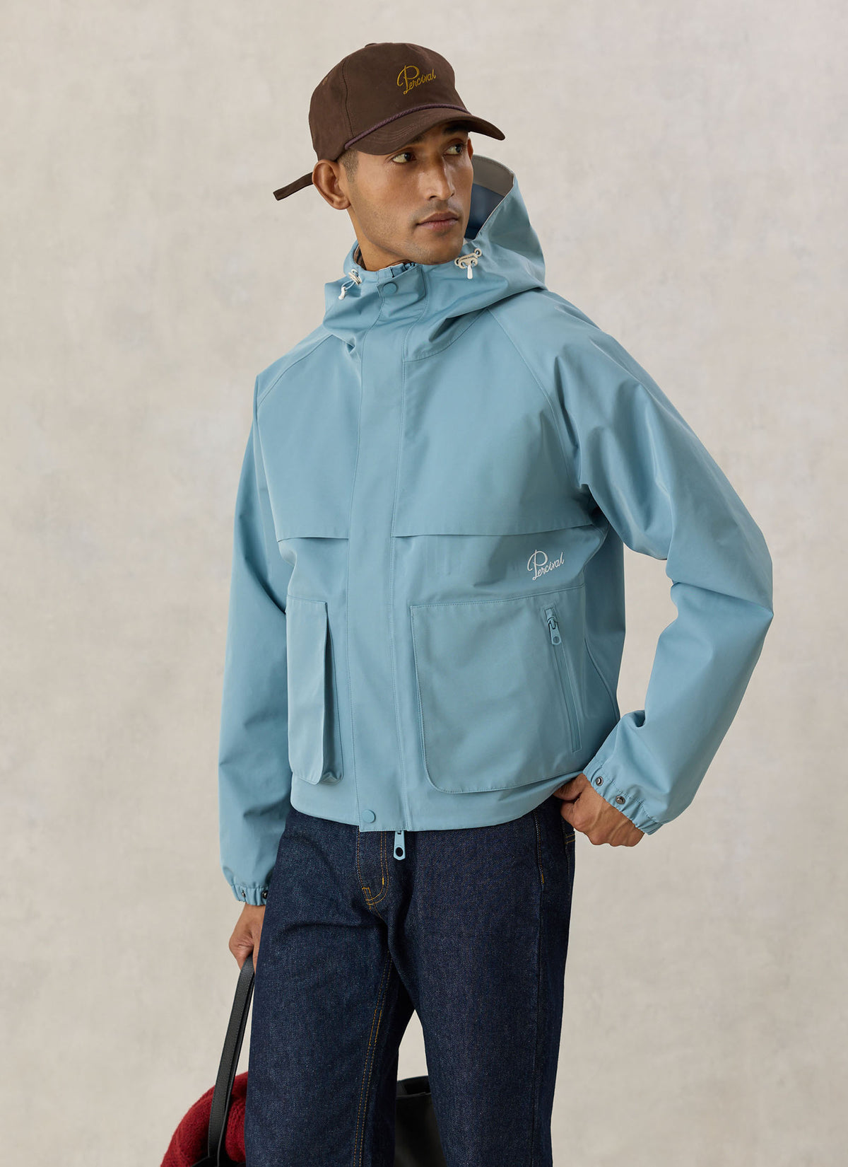 A model wearing the light blue Percival Hanson waterproof rain jacket with a boxy fit, paired with dark jeans and a cap.