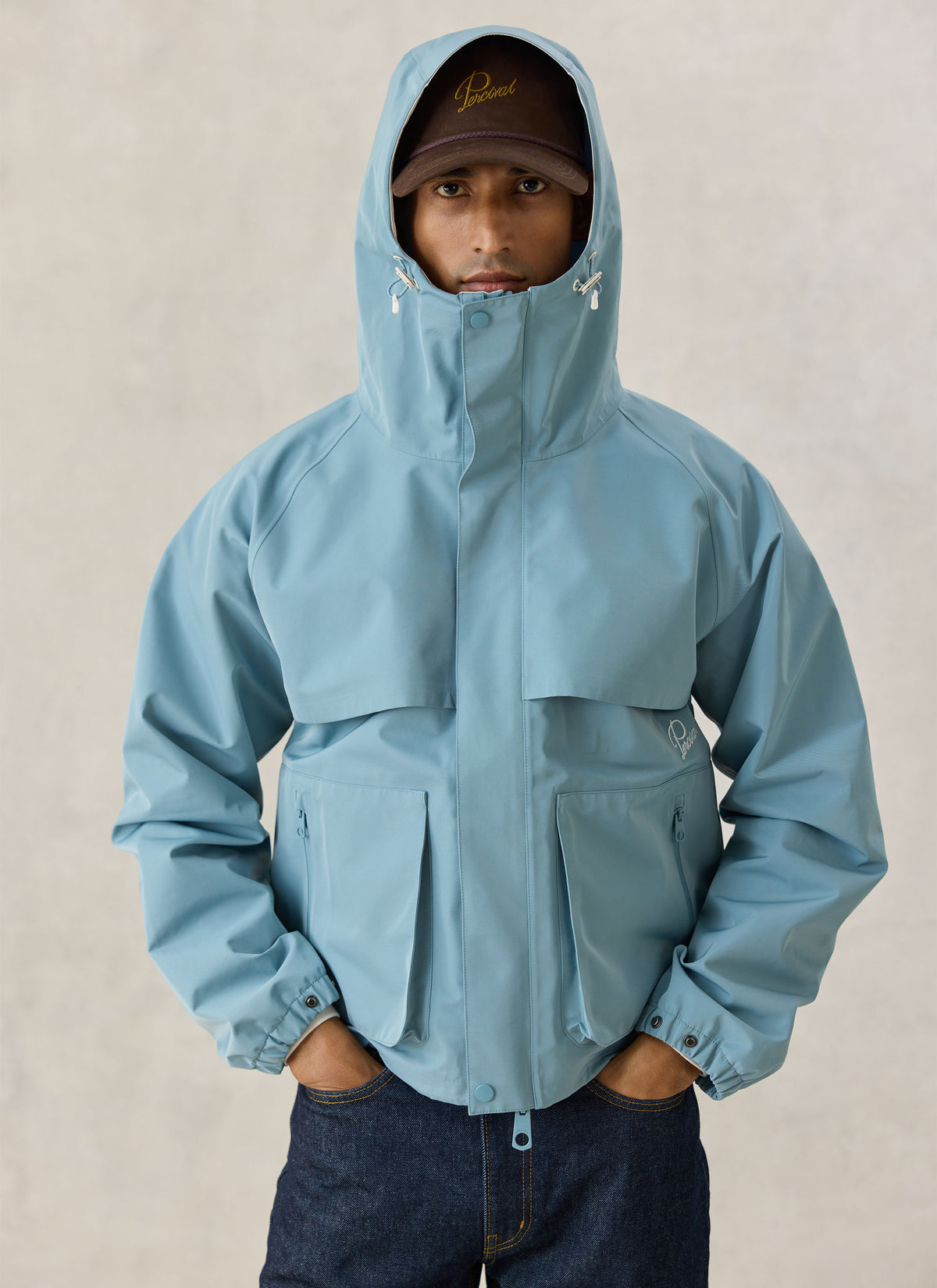 A model wearing the Percival Hanson waterproof rain jacket in light blue with the hood up against a neutral background.
